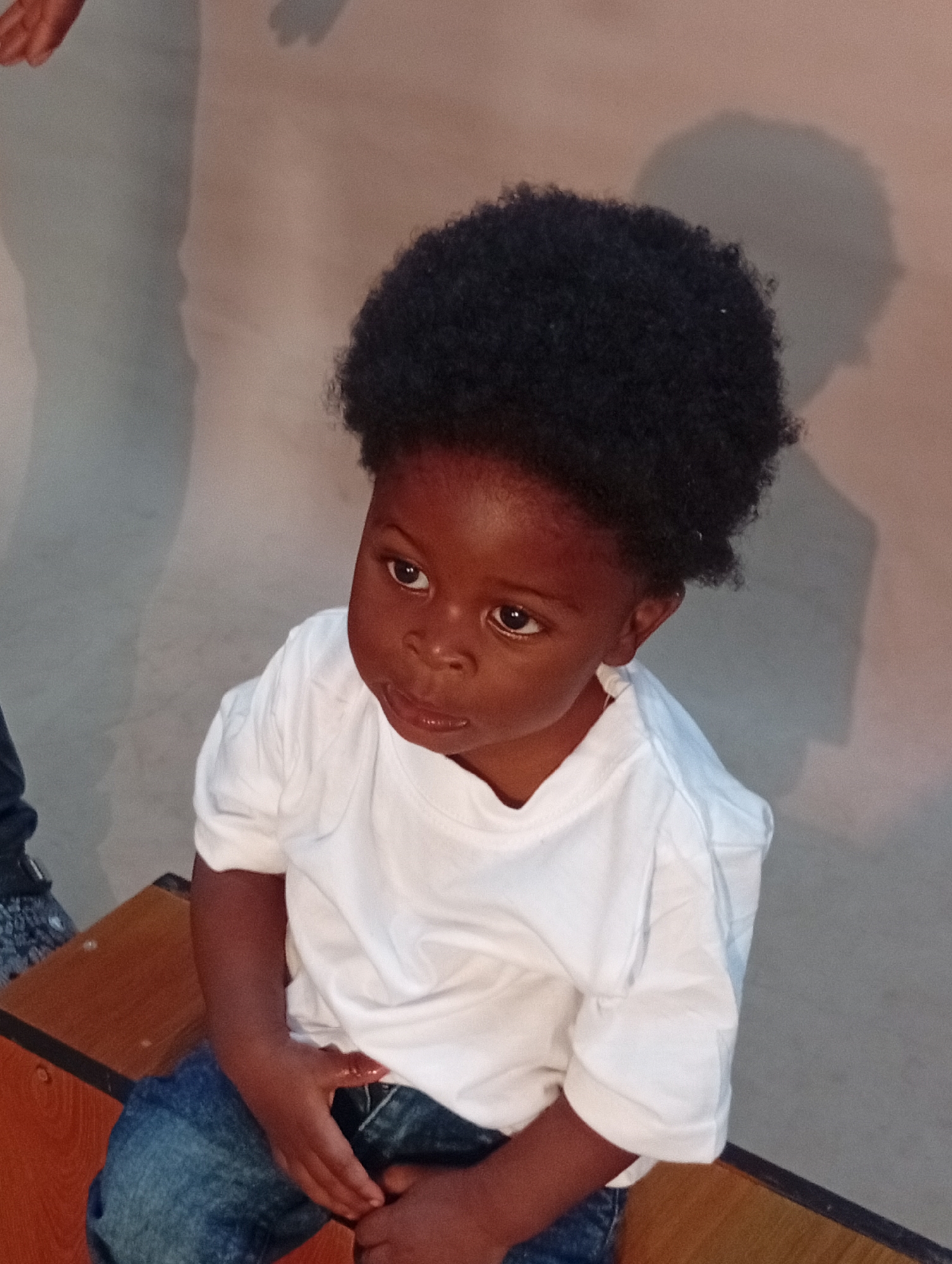 A young Black child with a full afro hairstyle is sitting and looking to their right. The child is wearing a plain white t-shirt and blue denim jeans. They are seated on a wooden bench with a dark border. The background is a wall that appears to be painted in a gradient from a light grey on the left to a light peach color on the right. Shadows of people are visible on the left side of the wall. The overall lighting suggests an indoor setting, possibly a community center or a similar public space.