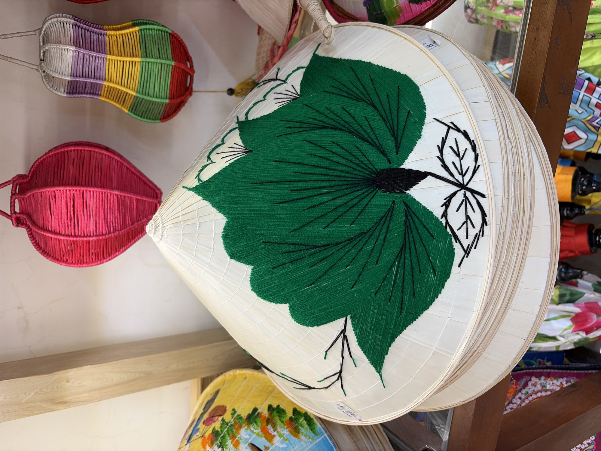 In a market stall in Da Nang, Vietnam, a stack of conical hats, known as "nón lá", is displayed. The top hat, prominently featured, is made of natural straw and adorned with a large, embroidered green leaf motif. Several other nón lá are stacked beneath it, with the top visible hat having a price tag attached. In the background, a wooden beam bisects the frame, and two decorative woven lanterns, one bright pink and the other multi-colored, hang from above. Other market items, including fabric with floral patterns and more lanterns, are visible in the lower portion of the image. The overall impression is one of a vibrant, local market setting.