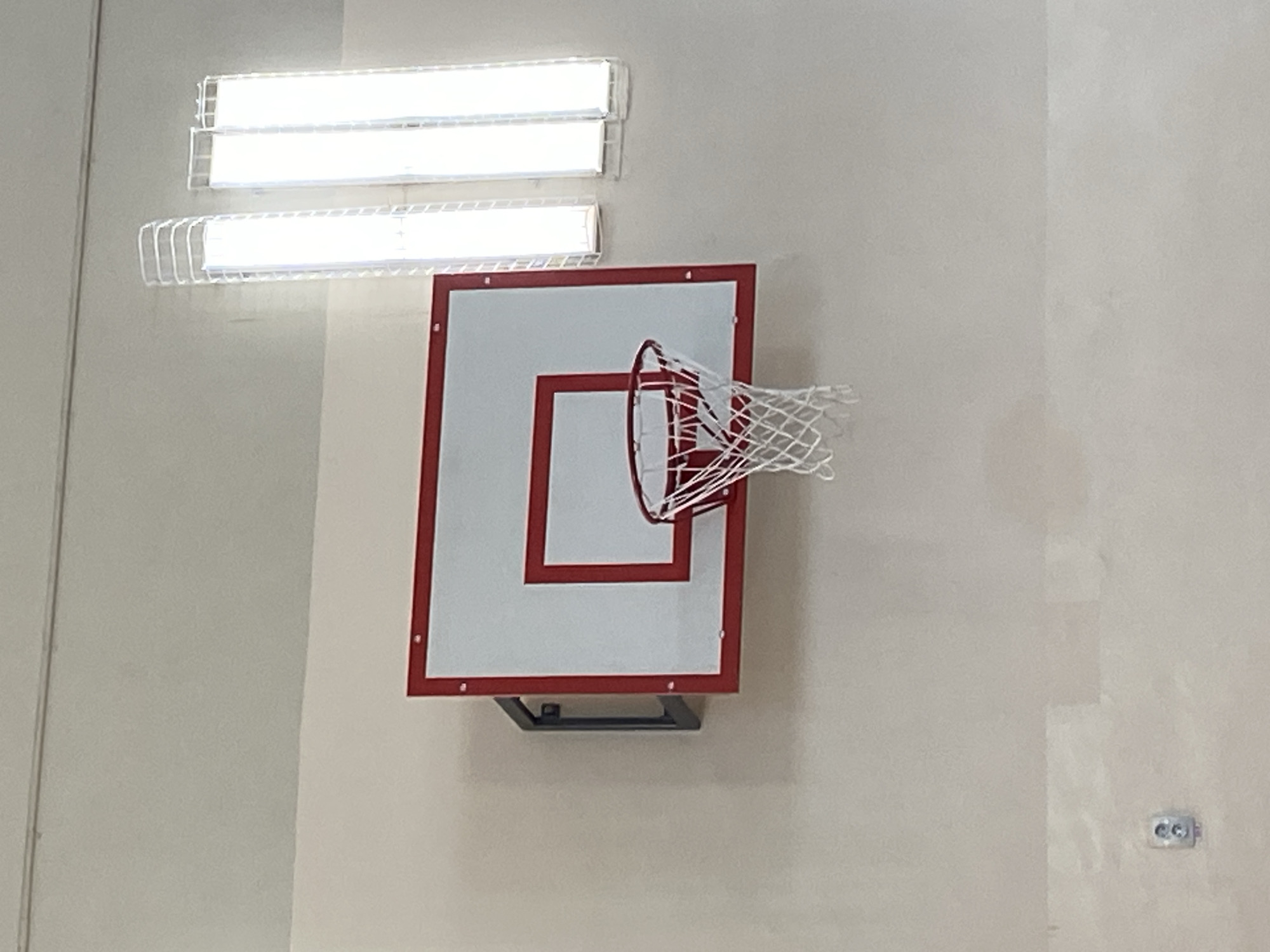 A basketball hoop and backboard are mounted on a wall in an indoor gymnasium. The backboard is white with a red border and rim. The net is white and appears to be made of string. In the background, there are two long fluorescent light fixtures. On the lower left of the image, there is a standard electrical outlet on the wall. The lighting suggests the image was taken indoors, possibly in a school or community center. The provided information states the image is from Votkinsk, Russia, but there are no visual cues within the image to confirm this specific location.
