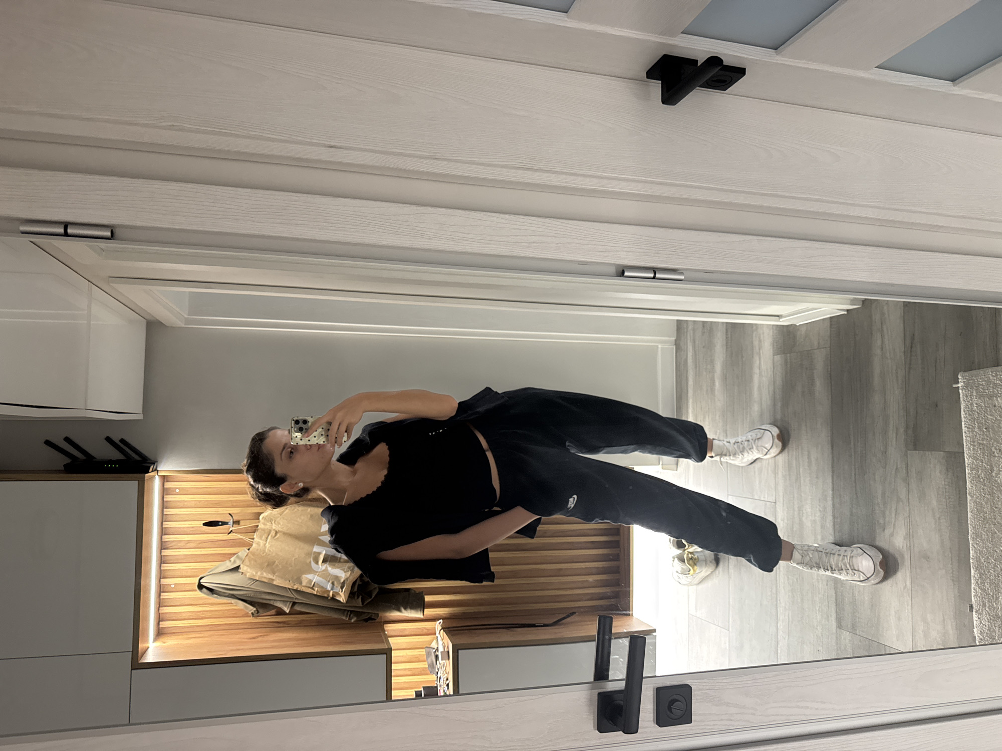 A woman in a black top and black sweatpants stands in a room. She is holding a cell phone in her left hand, seemingly taking a photo of herself in a mirror. The room has light grey wood-look flooring and white walls. A cabinet with light grey doors and wooden detailing is visible in the background. An unseen light source illuminates the wooden detailing, creating a warm glow. A black router with antennae sits on top of the cabinet. A jacket or bag is draped over the cabinet. The reflection of the room and the woman is visible in a mirror framed by a white door. Black door handles and hinges are on the door frame. The location is unspecified, and there is no indication that it is Burgas, Bulgaria.