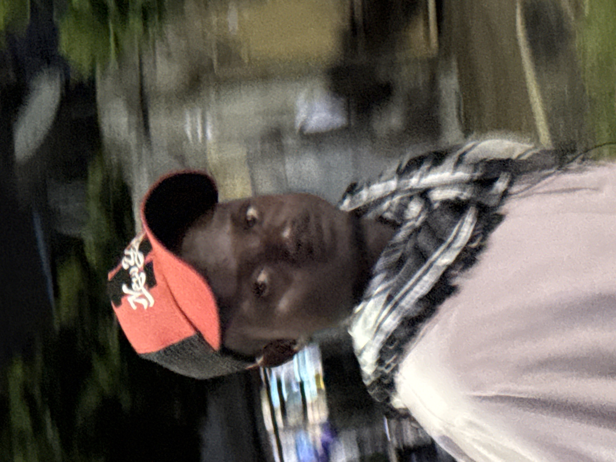 The image shows a medium shot of a young Black male wearing a red and grey baseball cap with the word "New York" written in white script. He is also wearing a light pink shirt and a checkered scarf around his neck. The man has dark skin, dark eyes, and a mustache. He is looking towards the camera with a neutral expression. The background is blurred, suggesting motion, and consists of dark trees and some indistinct structures. The lighting is dim, with some artificial light sources visible in the background. The image appears to have been taken at night or in low-light conditions, possibly outdoors in an urban or suburban environment. There are no obvious landmarks to pinpoint the location within Abuja, Nigeria.