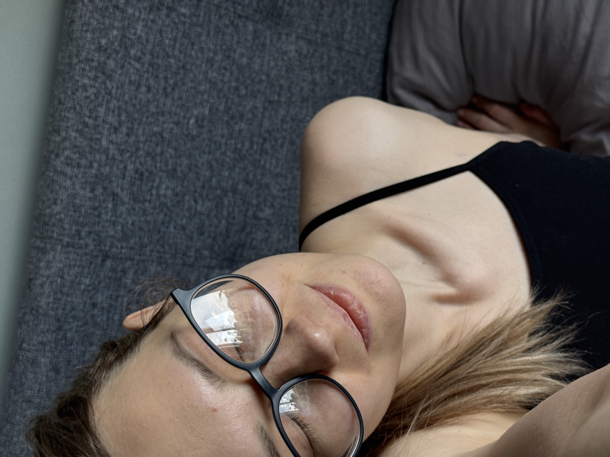 This is a high-angle, tilted selfie of a person, likely female, lying down indoors. The subject is seen from the chest up, with her head resting on a surface. She has light brown or blonde hair, wears black-rimmed glasses, and a black camisole or tank top. Her eyes appear closed or are looking downwards, and her mouth is slightly open, conveying a relaxed or pensive expression. Her skin is fair, with some freckles visible.
The background primarily consists of a textured grey fabric surface, likely a couch or bed, on which the person is resting. To the upper right, a dark grey or brown pillow is visible, with what appears to be a hand partially obscured behind it. The lighting suggests an indoor setting, possibly during the day or with artificial light. No specific activity beyond resting or taking a selfie is clearly depicted. There is no visible text in the image. The location context provided is Chaikovsky, Russia.