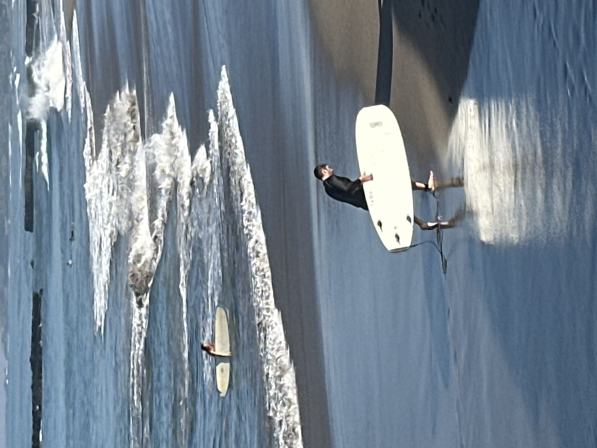 In North Kuta, Indonesia, on a beach, a man wearing a black wetsuit carries a white surfboard. He is walking from the water onto the shore, with the surfboard held horizontally. In the ocean, another surfer, partially visible, sits on a white surfboard. The waves are breaking and creating white foam. The water is blue and reflects the sky. The sand on the beach is dark and wet from the tide.