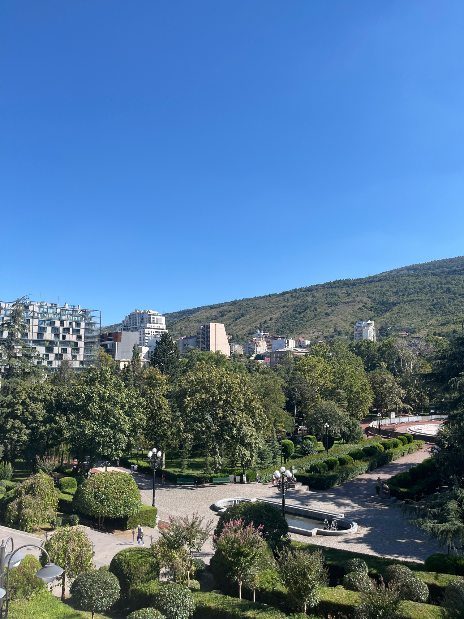 The image depicts an elevated view of a city park in Tbilisi, Georgia, on a clear, sunny day. In the foreground, manicured bushes and trees line paved pathways. A small fountain with jets of water is visible. Further into the park, lampposts illuminate the walking paths, and several people are seen strolling. Beyond the park, a row of modern and older buildings rises, including a distinctive building with a glass facade. In the background, a lush green, tree-covered hill forms the natural landscape, with some structures dotting its slopes. The sky above is a brilliant, cloudless blue. The overall scene conveys a sense of urban tranquility amidst natural beauty.