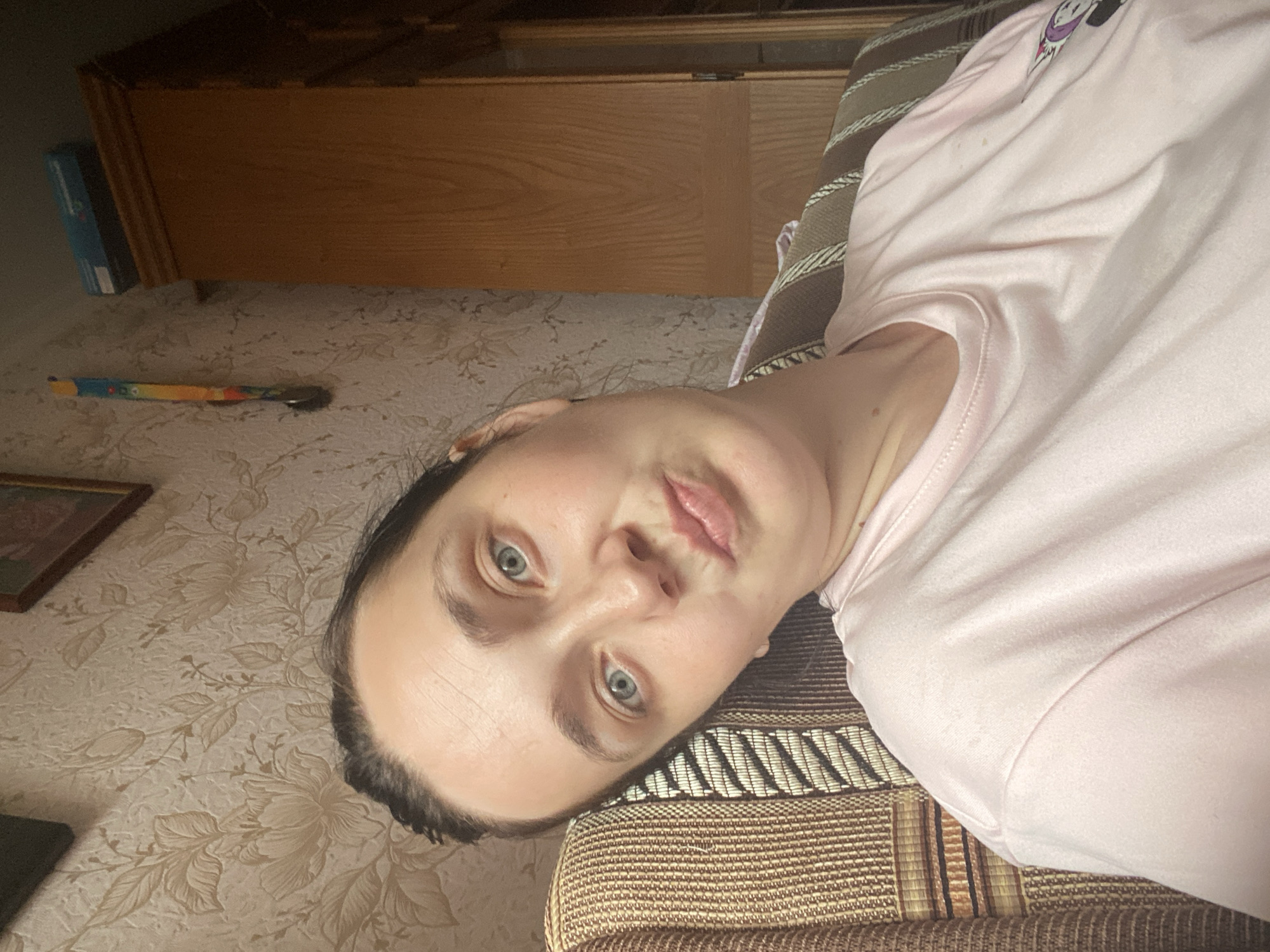 A young woman with light blue eyes and dark hair pulled back is looking at the camera with her head tilted to the side, leaning against a striped chair. She is wearing a pale pink t-shirt. She is indoors, in what appears to be a living room, with patterned wallpaper on the wall behind her. A wooden cabinet is visible to her right, and a framed picture hangs on the wall above her.