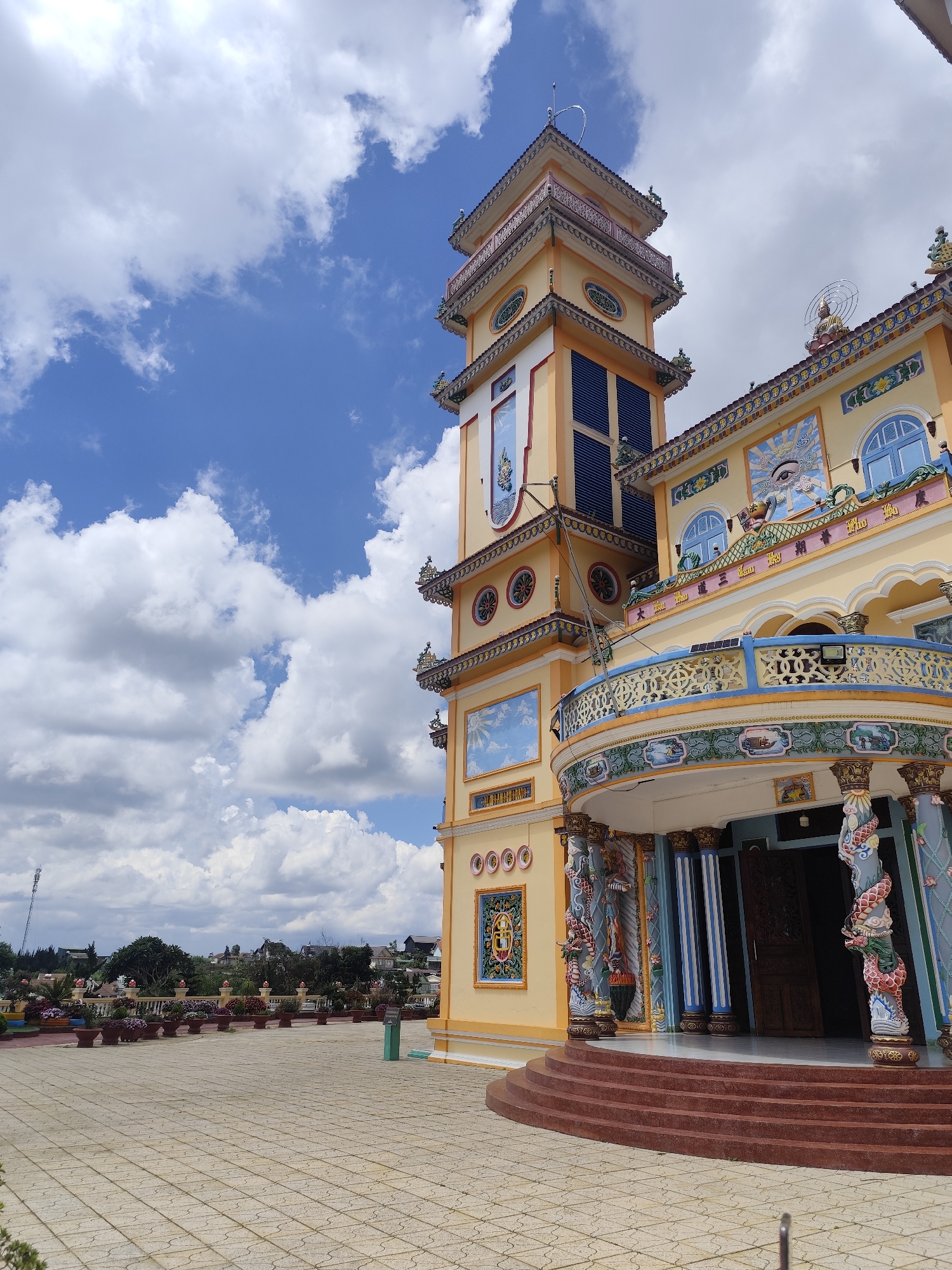 The image shows the exterior of a vibrant yellow Buddhist temple with ornate architectural details, located in Xuân Thành, Vietnam. The temple features a tall, multi-tiered tower with decorative elements and paintings, and a curved balcony adorned with intricate blue and gold patterns. The entrance to the temple is marked by a set of red stairs and blue and white striped columns, with elaborately decorated pillars depicting dragons. The temple is set against a backdrop of a bright blue sky with fluffy white clouds. In the foreground, a paved courtyard leads to the temple, with a row of potted plants and trees lining the perimeter in the middle ground. Beyond the temple grounds, a residential area with houses and buildings is visible, nestled amongst trees and hills. The scene is brightly lit, suggesting it was taken during the daytime. There are no people visible in the image.
