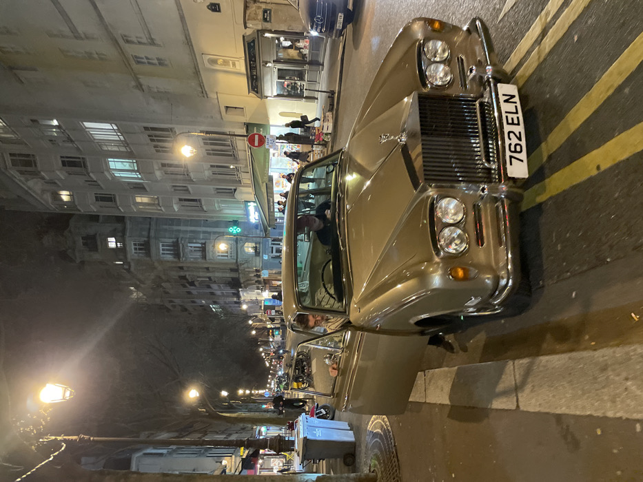 The image depicts a nighttime street scene in Paris, France, specifically on Rue Monsieur le Prince in the 6th arrondissement. A vintage, gold-colored Rolls-Royce Silver Shadow, bearing the front license plate "762 ELN," is prominently positioned on the asphalt street, partially over double yellow lines.
Inside the vehicle, a male driver, wearing a dark cap, is visible behind the steering wheel. The front passenger door is open, and a male individual wearing glasses and a striped shirt is in the process of exiting the vehicle, with his feet touching the pavement.
The street is illuminated by multiple ornate streetlights lining both sides. On the left, a paved sidewalk features a tree grate and a grey wheeled refuse bin. On the right, the sidewalk borders multi-story Parisian buildings with classic architecture, some windows illuminated. Ground-level shops are visible, including one with a green awning and a prominent luminous green cross, indicating a pharmacy. Another shop displays a sign reading "Meillet." A red circular "No Entry" sign is also visible above one of the shops.
Several pedestrians are present on the sidewalks in the background. Further down the street, a blue car (license plate "GN-221-HN" visible) is parked on the right, and a scooter is parked on the left.