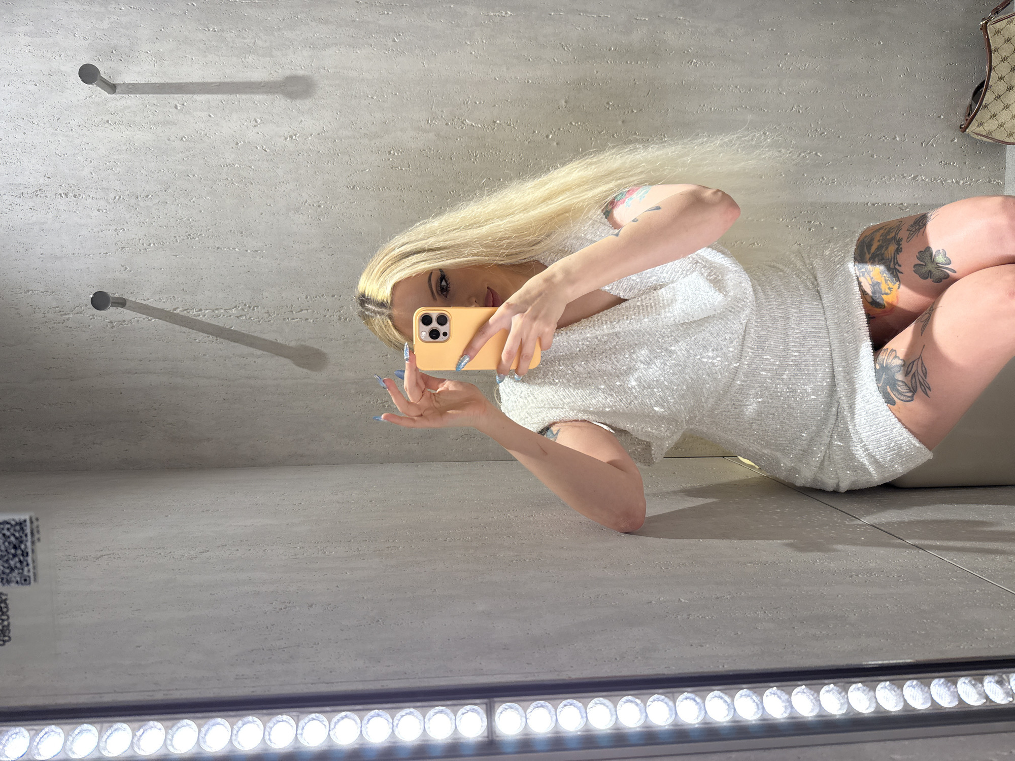 The image shows a woman with long blonde hair taking a mirror selfie. She is wearing a sparkly white dress and has tattoos on her arms and legs. She is holding a light orange smartphone to take the picture. In the background, there are two metal pegs on a textured wall and a handbag is visible on the right side of the image. The floor appears to be tiled and there is a row of bright lights at the bottom of the frame. The image is taken in a mirror, so the woman is facing away from the viewer, looking at her reflection. The lighting suggests it could be daytime, but the indoor setting makes it difficult to determine. The overall mood is casual and confident. There is a QR code visible on the left side of the image.