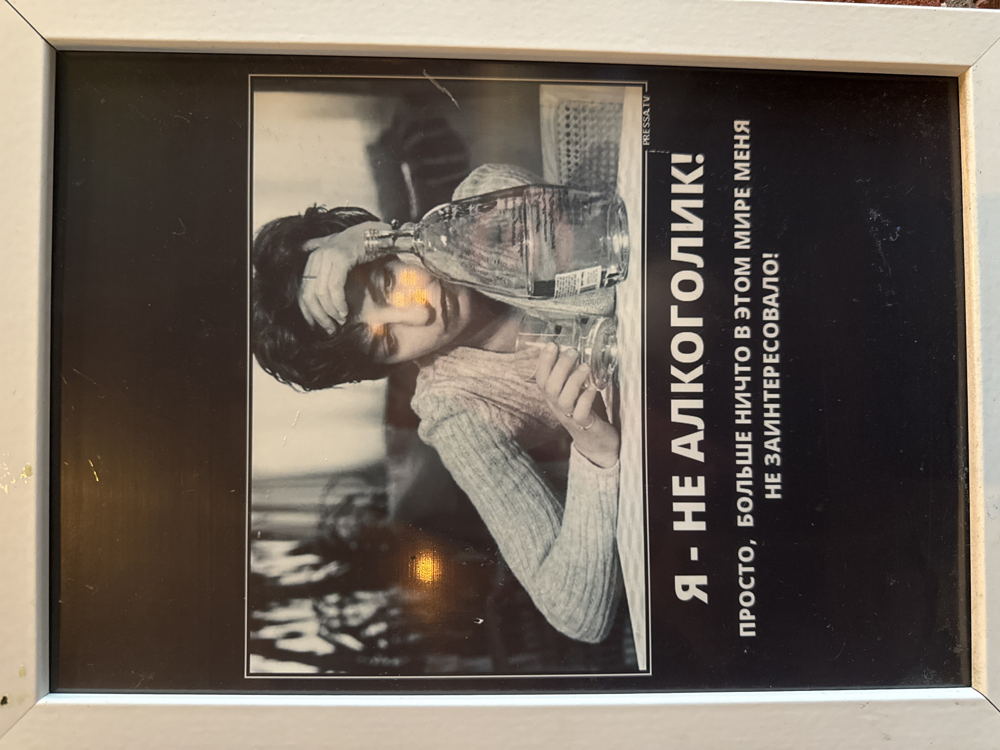 The image shows a framed poster in black and white. The poster features a person, possibly male, leaning with their head on a tilted glass bottle, presumably an alcoholic beverage. The person is wearing a light-colored, ribbed sweater. A glass rests beside the bottle. The poster includes Russian text in bold, which, according to OCR, states "I am not an alcoholic! Simply, nothing else in this world has interested me!" The top right of the poster has the name "PRESSA.TV". The frame around the poster is white, and the background appears to be indoors. There is no definitive information to confirm it is Moscow, Russia.