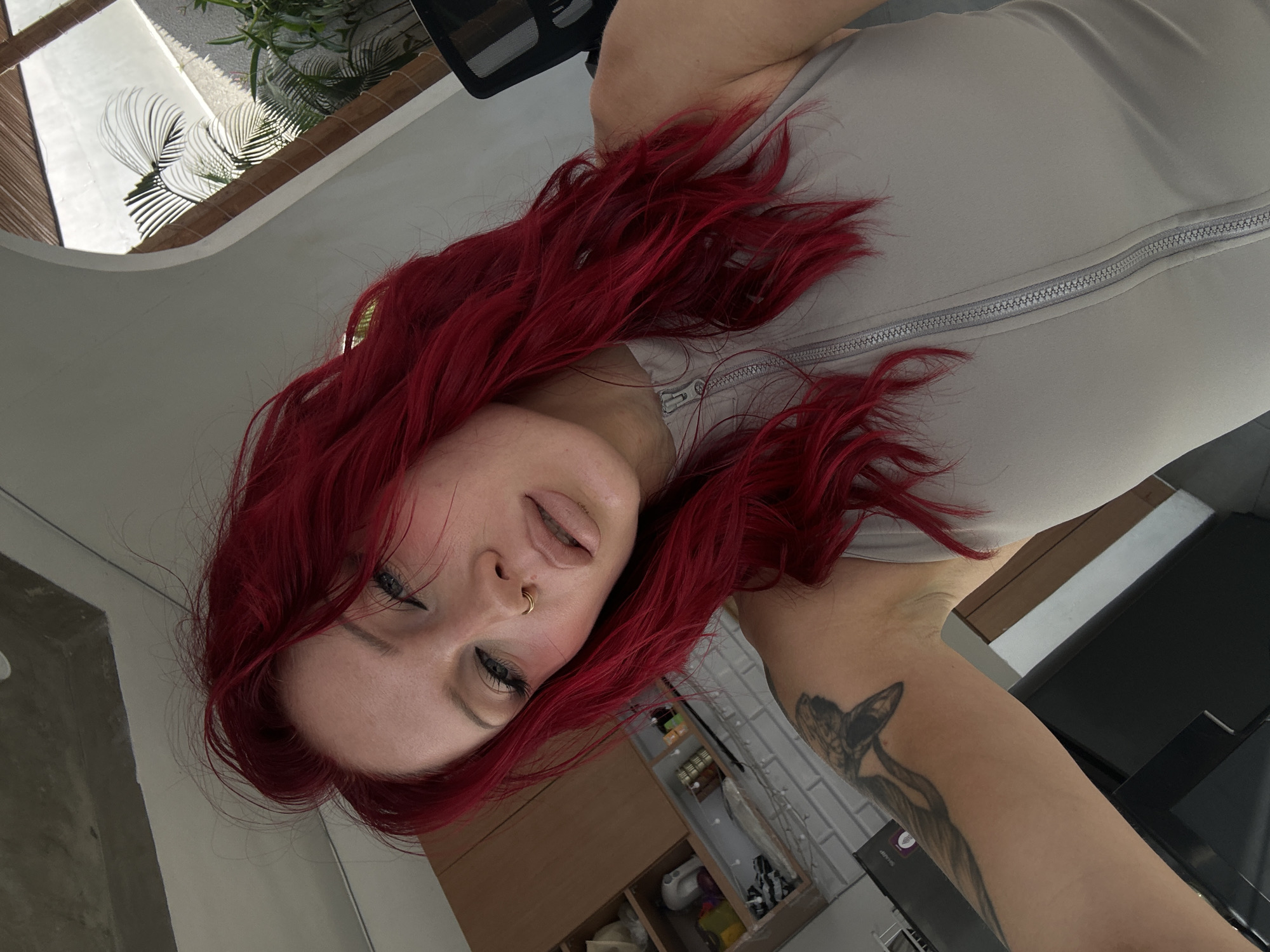 A selfie shows a woman with bright red hair posing in what appears to be a domestic setting. She is wearing a light gray, sleeveless zip-up top. Her face is turned towards the camera, with her left arm extended, showing a tattoo on her bicep. She has a nose ring and makeup, including eyeliner and blush.
The background reveals elements of a kitchen or living area with wooden cabinets, white brick-like tiling, and a gray ceiling. Through an arched opening, an outdoor scene is visible, with lush green plants and natural light suggesting a tropical environment, consistent with North Kuta, Indonesia.