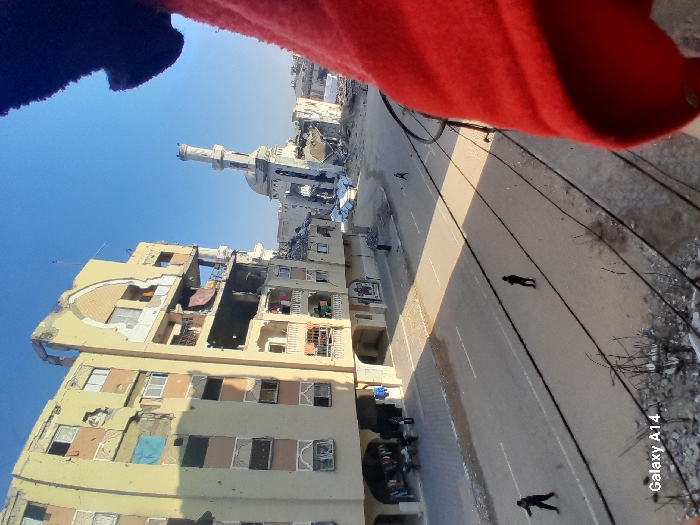 The image presents an elevated view of a street in Khan Yunis, Palestinian Territory, under a clear blue sky. On the left, a multi-story, light-colored building exhibits extensive structural damage, including large impact holes, missing facade sections, exposed interiors, and shattered windows; colorful fabrics hang from some openings. Below this building, on the sidewalk, several individuals are gathered near a ground-floor area displaying merchandise. Further down the street, a white mosque with a distinctive dome and a partially damaged minaret is visible, surrounded by significant amounts of rubble and severely damaged structures. The asphalt road in the foreground has white lane markings and is occupied by three individuals walking: one in dark clothing on the left lane, another in dark clothing on the right lane further back, and a third distant figure on the right lane. Overhead wires are visible in the lower foreground, and debris lies on the ground beneath the vantage point. A red fabric object partially obscures the right side of the frame.