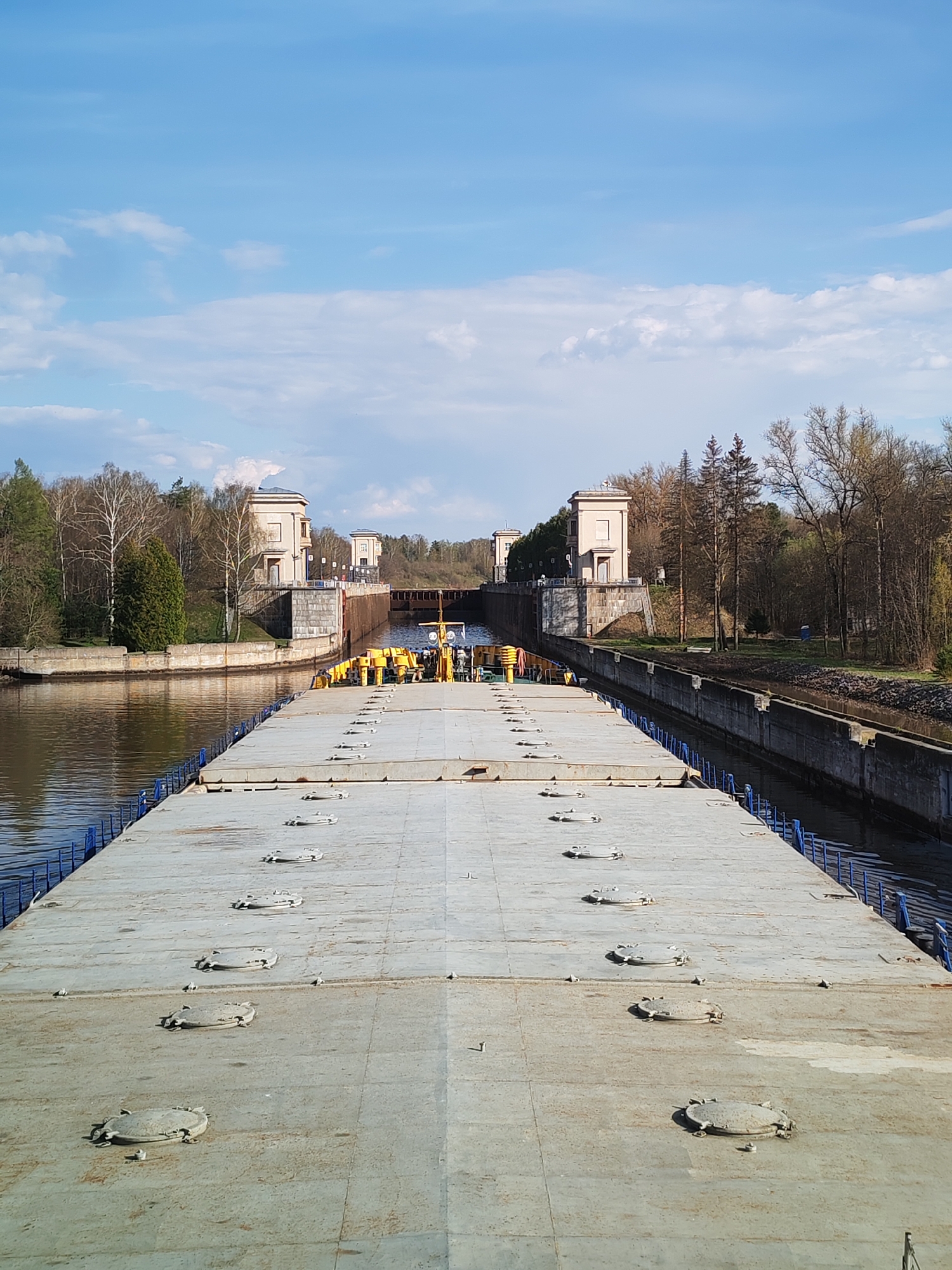 The image depicts a wide, flat, gray barge navigating a canal. The barge deck is lined with numerous circular access hatches. Blue railings run along the edges of the barge. The canal is bordered by concrete walls on both sides. Beyond the canal walls, trees and vegetation line the banks. In the distance, a series of buildings with architectural details reminiscent of classical design are visible on both sides of the canal. A metal pedestrian bridge spans the canal between the buildings. The sky is blue with scattered clouds. The location is Unknown, Unknown.