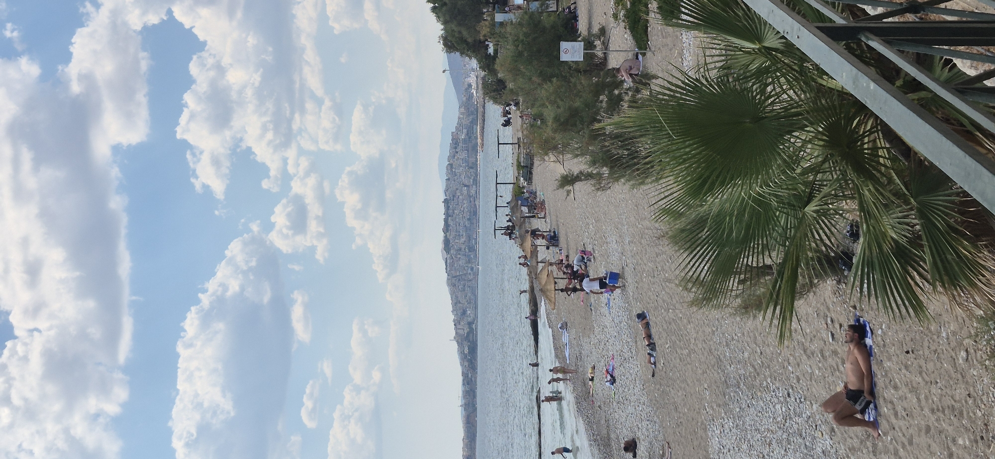 A vertical, medium shot captures a lively beach scene in Paleo Faliro, Greece, under a partly cloudy sky. The foreground is dominated by a lush green palm frond and a metal railing, hinting at a viewpoint from an elevated position. The sandy beach, dotted with pebbles, slopes down to the sea, where several people are wading and swimming. Further down the beach, numerous sunbathers are scattered across towels and mats, some seeking shade under thatched umbrellas. In the distance, a picturesque city skyline stretches across the horizon, nestled against rolling hills. A boat is visible sailing in the calm waters. The overall atmosphere is one of a relaxed summer day at the seaside.