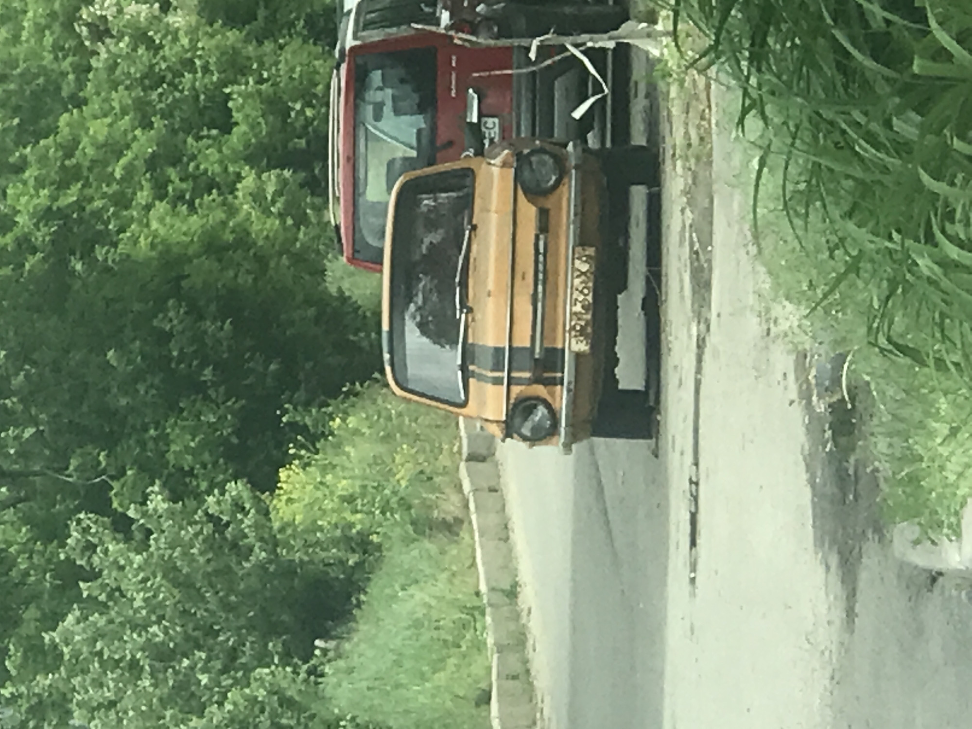 The image shows a street scene in an unknown city. A yellow ZAZ-968 car with black stripes is parked on the side of the road. Behind it, partially visible, is a red vehicle, possibly a truck. The environment consists of greenery, including trees and grass, on the left side of the road. The road is paved and appears to be in an area that might be semi-rural or on the outskirts of a city, as indicated by the presence of trees and foliage. No people are visible in the image. The location and time of the photograph are unspecified.