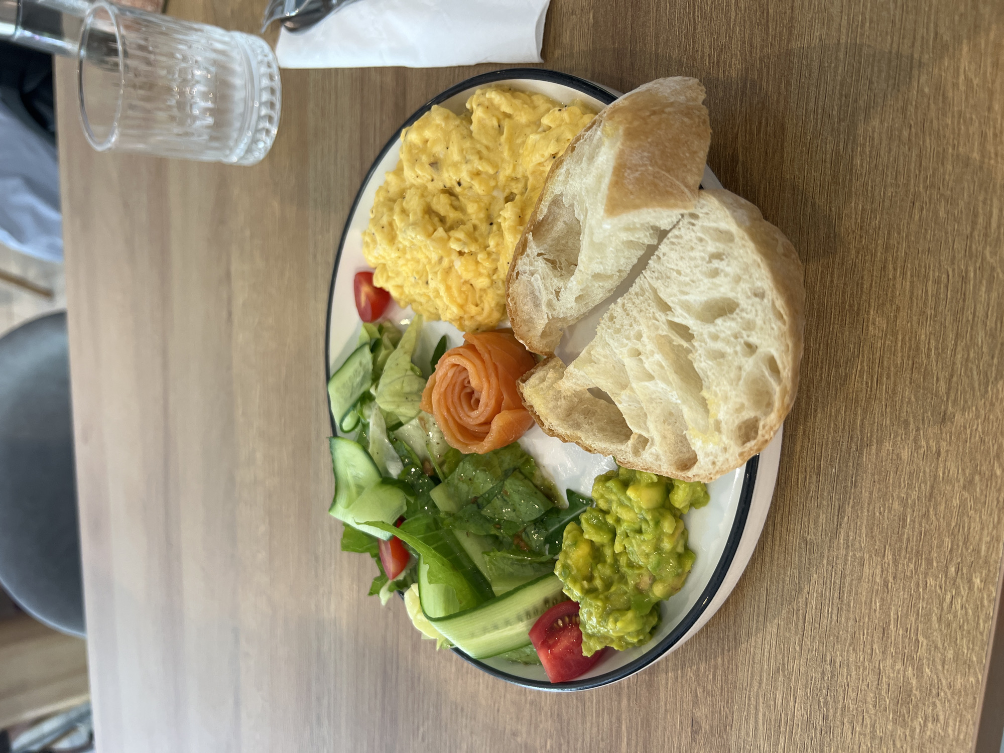 A plate of food is centered on a light-brown wooden table. The plate contains scrambled eggs, bread, a salad of lettuce, cucumbers, and tomatoes, avocado, and salmon. There is also a glass and utensils in the background. The location is in Nha Trang Ward, Vietnam.