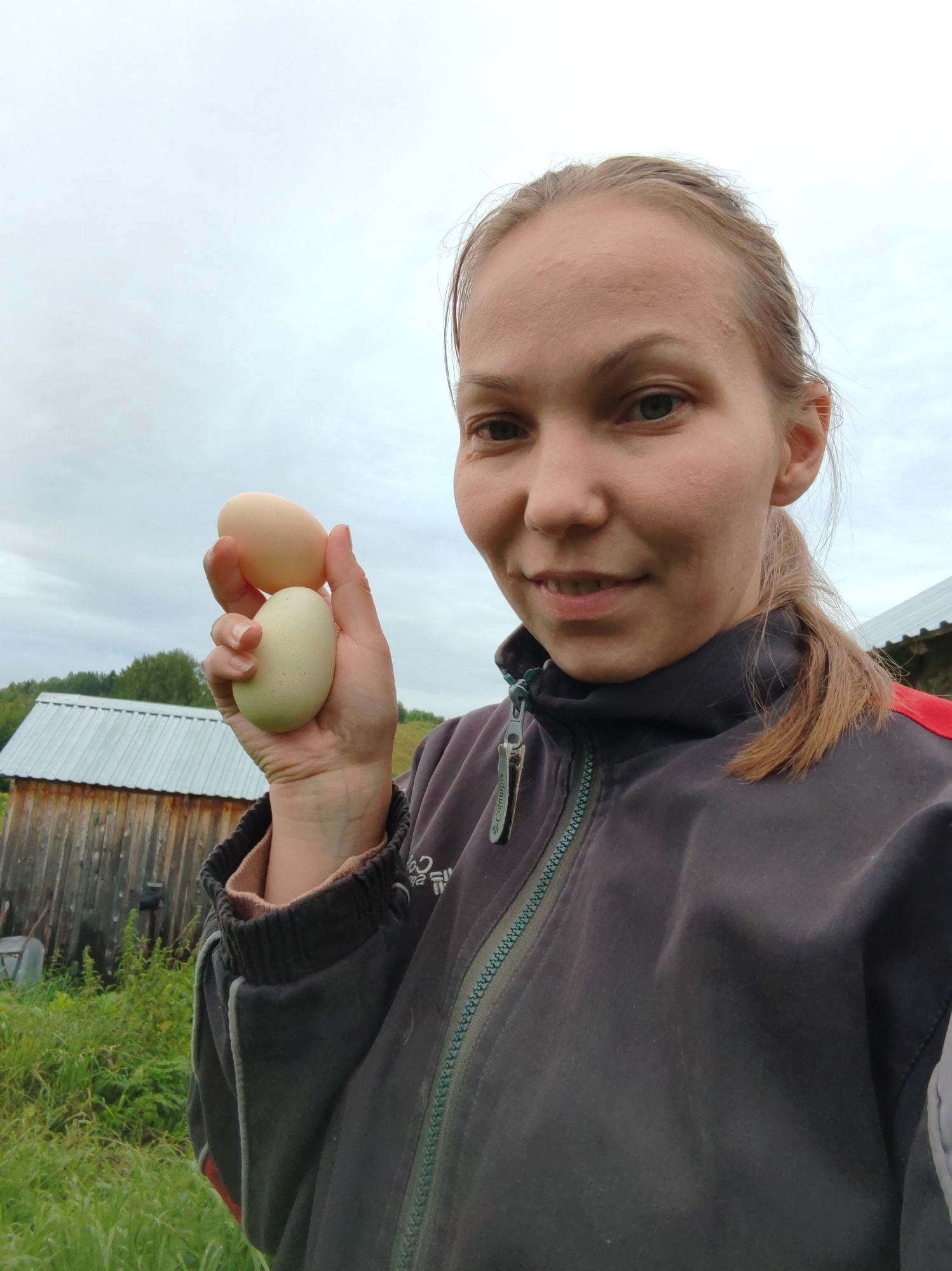 A close-up, eye-level, outdoor, selfie-style shot shows a woman in a grey jacket holding two eggs in her right hand, which is extended towards the camera. The woman's head and upper torso are in focus, with her face turned slightly towards the camera. She has blonde hair pulled back in a ponytail and is looking directly at the viewer with a subtle smile. The eggs appear to be light brown and pale green.
Behind her, a rustic wooden structure with a corrugated metal roof is visible on the left side of the frame, partially obscured by green foliage. The background consists of a cloudy sky and a distant line of trees, suggesting a rural or semi-rural setting. There are no specific landmarks visible that would indicate the city of Чухлэм, Russia, or any other specific urban location. The overall impression is that of someone in a farm or outdoor environment.