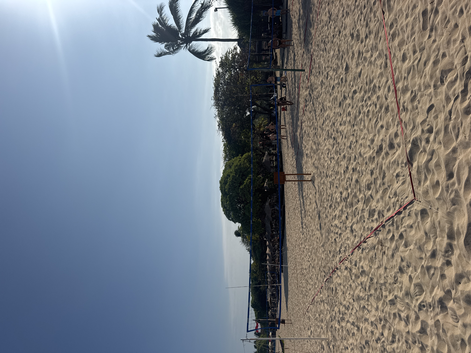 The image depicts a sandy beach volleyball court in Kuta Selatan, Indonesia, bathed in bright sunlight. A tall volleyball net stretches across the court, with red boundary lines visible on the sand. Several people are scattered on the court, some appearing to be playing or preparing to play volleyball, while others are standing or walking around. In the background, a line of thatched-roof umbrellas shades lounge chairs, suggesting a resort or beach club. Lush green trees form a dense backdrop, with a prominent palm tree swaying in the distance. The sky is a clear, bright blue, with a sun glare visible in the upper right corner, indicating a sunny day.