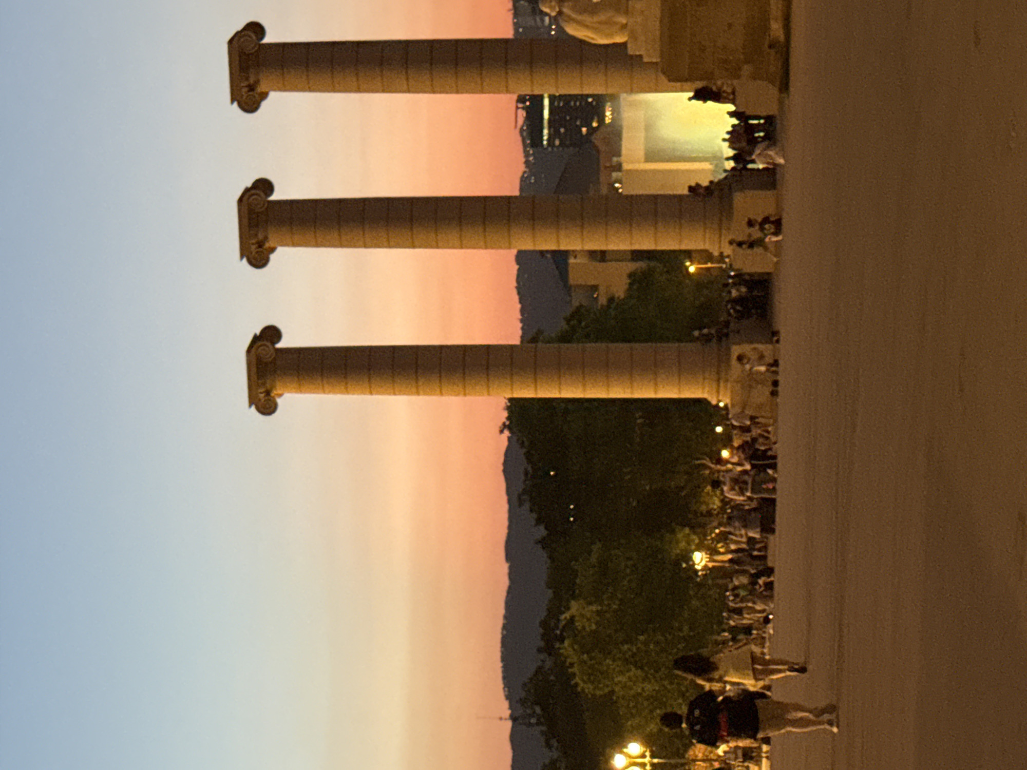 The image depicts the Four Columns of Montjuïc in Barcelona, Spain, at dusk. Three large, fluted stone columns with Ionic capitals stand prominently in the foreground, bathed in the warm glow of artificial lighting. The sky transitions from a pale blue at the top to a soft orange and pink gradient near the horizon, indicating sunset. In the middle ground, a crowd of people is visible, some walking, others gathered, near trees and streetlights. A statue, possibly of a nude figure, is situated to the right of the columns. The ground in the foreground is a flat, paved surface. The overall atmosphere is serene and public, with people enjoying the late evening in an open urban space.