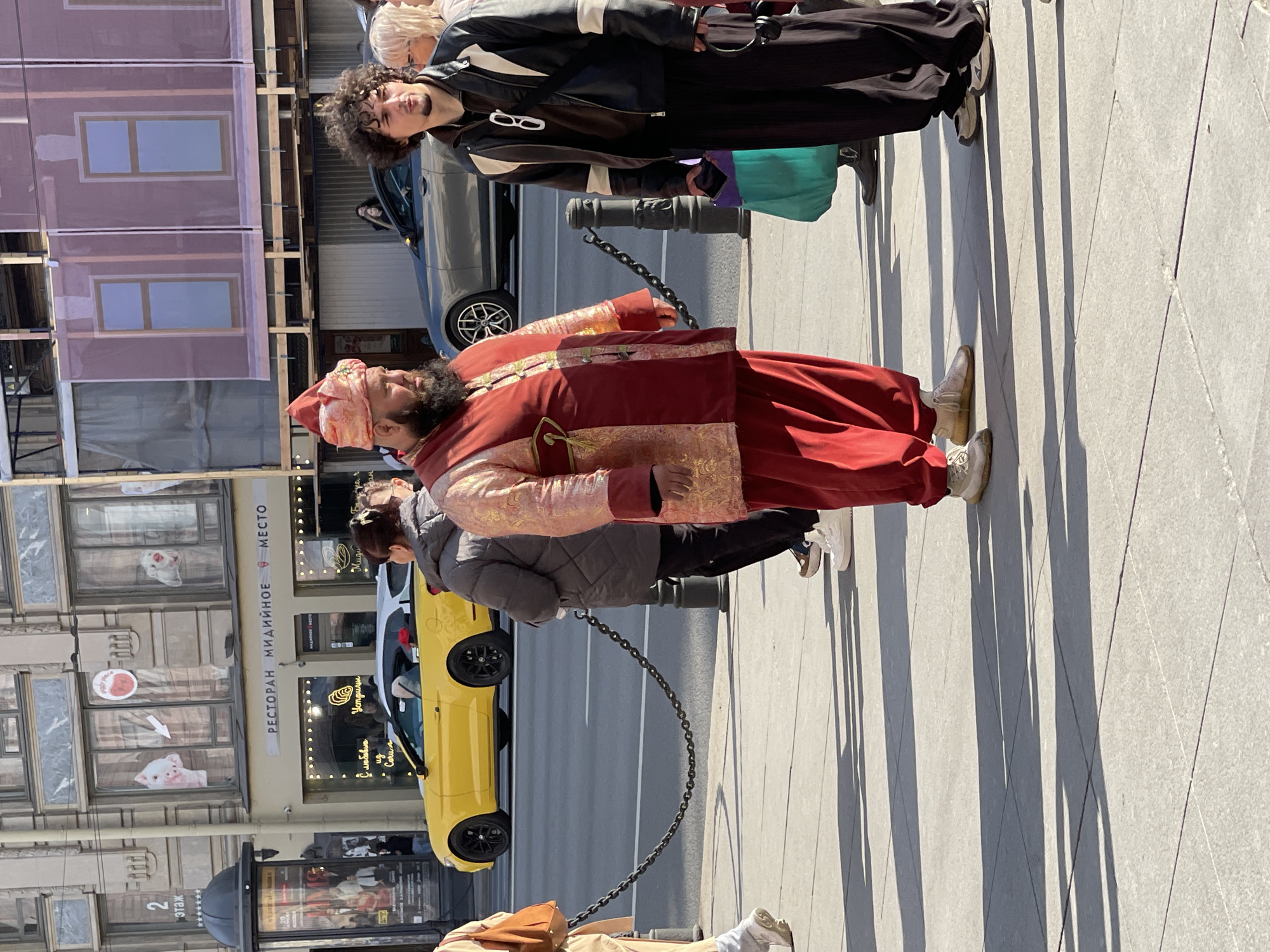 The image shows a street scene in Saint Petersburg, Russia. The main subject is a man dressed in a red and gold traditional robe and a red turban, standing on a sidewalk. There are several other people in the background, including a man in a black leather jacket, a person in a grey jacket, and another individual near the bottom of the frame. A yellow car and a grey car are visible on the street. A building with restaurant signage in Russian, "РЕСТОРАН МИДИЙНОЕ МЕСТО," can be seen behind the man in the robe. The sidewalk is made of grey tiles and has long shadows cast upon it. There are black posts connected by chains lining the sidewalk.