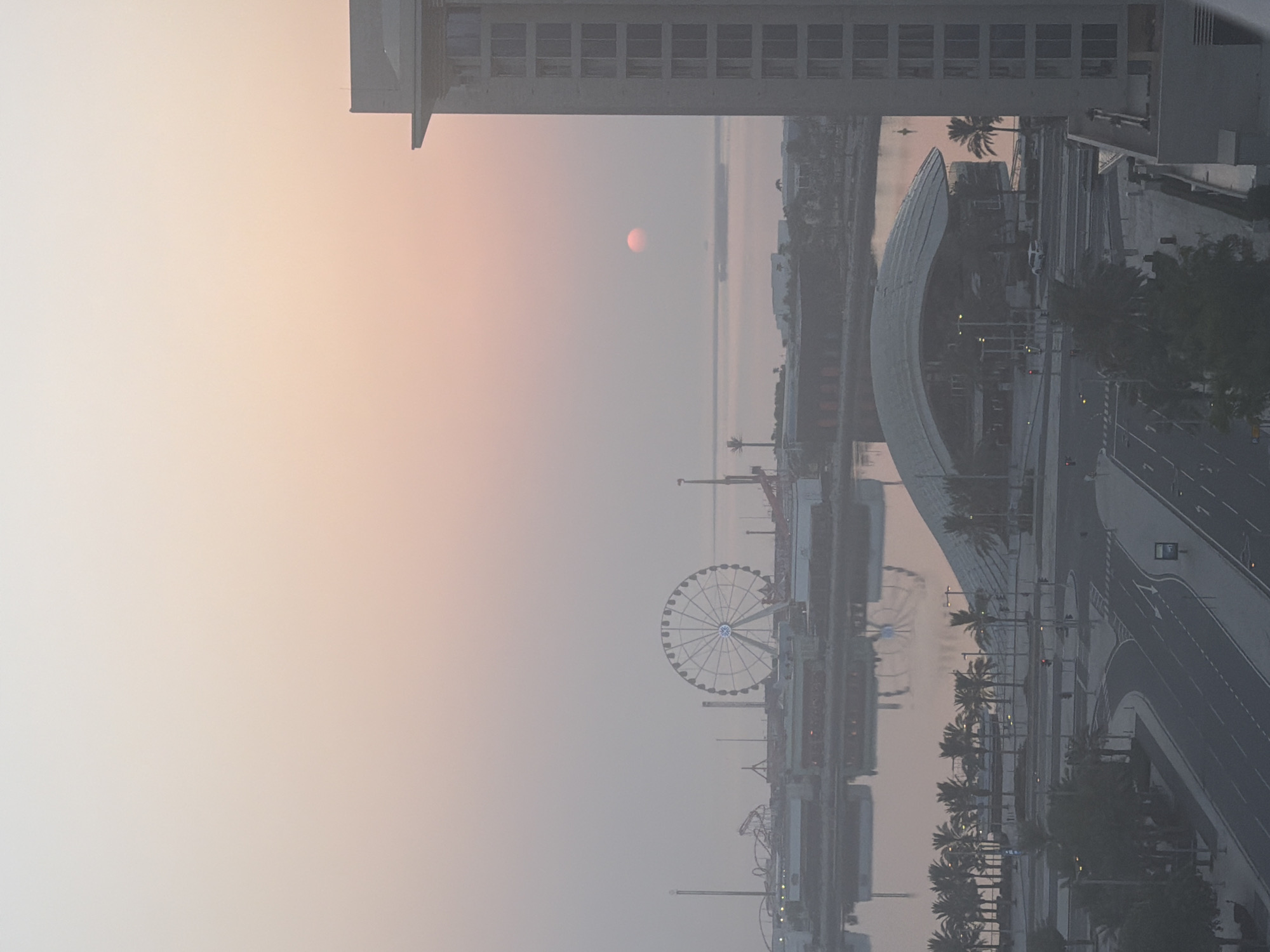 The image captures a hazy, pink-tinged sunrise over the city of Lusail, Qatar. In the foreground, a tall, modern building with a textured facade stands prominently on the right. Below this building, a street with multiple lanes and traffic signals is visible, lined with palm trees and other greenery. Across the water, a waterfront area features buildings, a distinctive modern structure with a curved roof, and a large Ferris wheel. In the distance, the sun appears as a small, reddish-orange orb low in the sky, casting a soft glow over the calm water and the hazy cityscape. The overall atmosphere is serene and somewhat muted due to the atmospheric conditions.