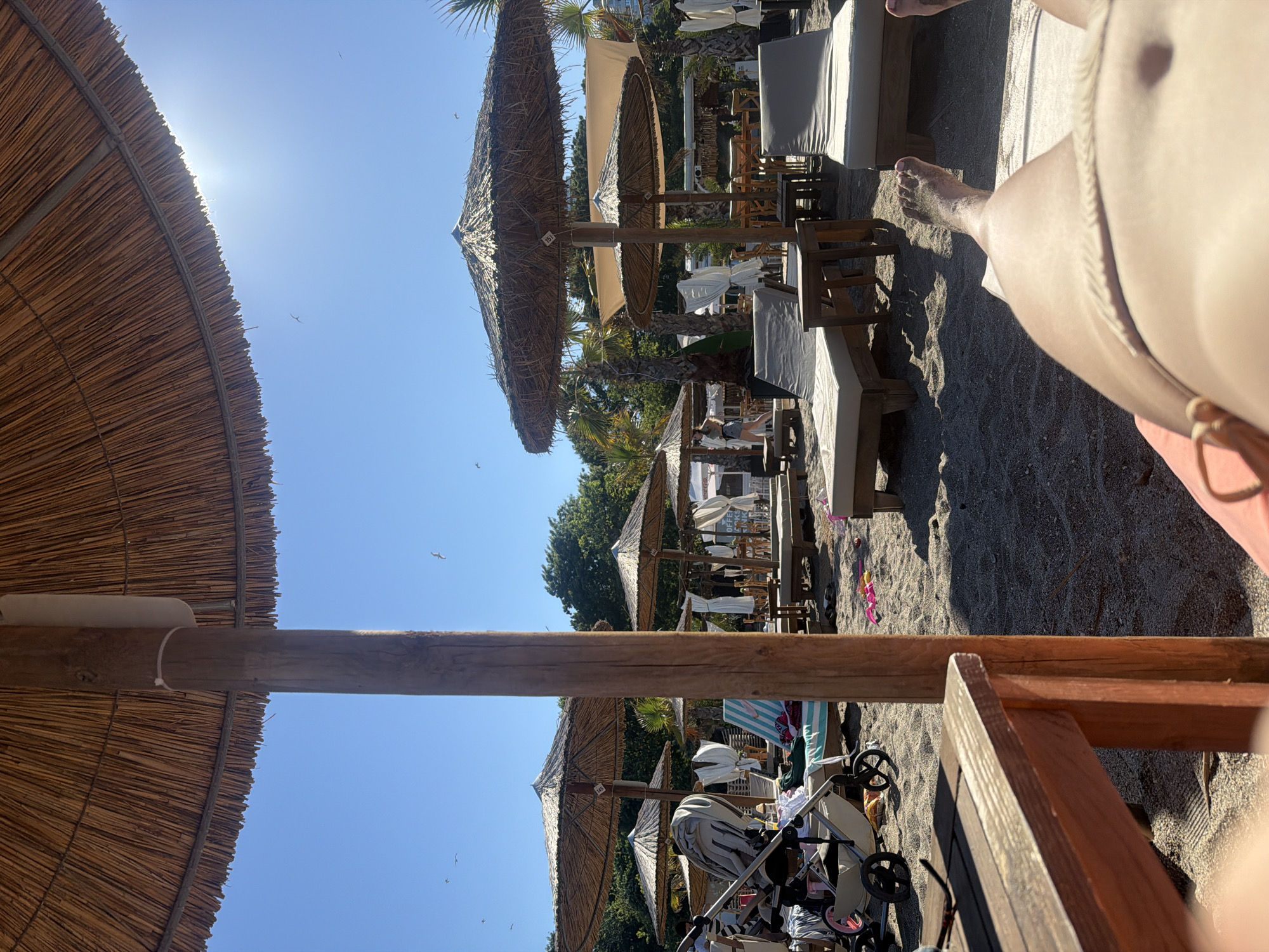This is a photograph taken on a sunny day at a beach in Burgas, Bulgaria. The perspective is from someone lying on a lounge chair, looking towards the sea. In the foreground, a wooden table is visible. Beyond that, the sandy beach is dotted with several thatched umbrellas and lounge chairs, some occupied by people. In the background, there are trees and a clear blue sky with a few birds flying. The scene suggests a relaxing day at the beach, likely a resort area given the amenities.