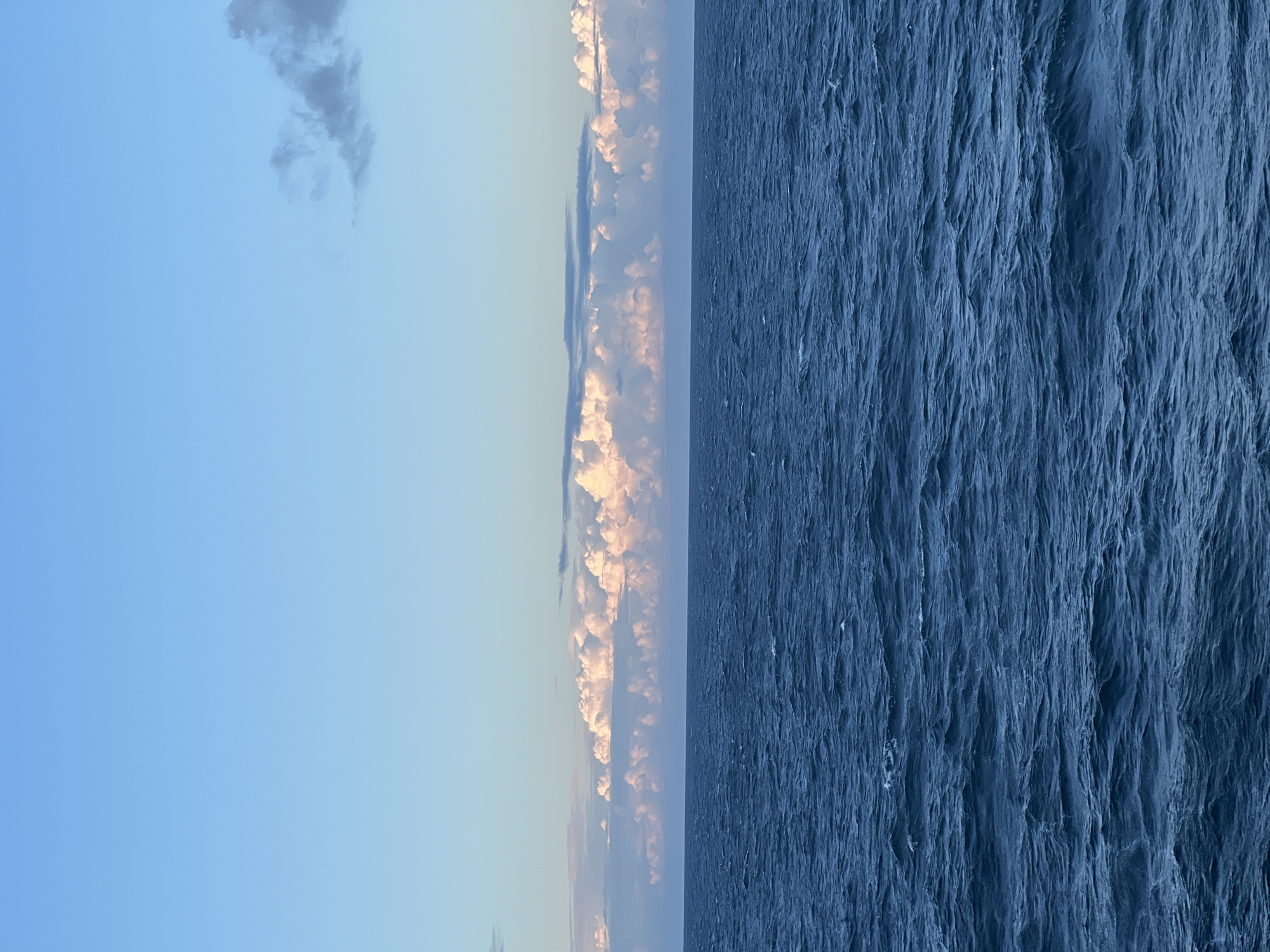 The image shows a vast expanse of deep blue ocean under a light blue sky. A line of fluffy, pink-tinged clouds stretches across the horizon, illuminated by what appears to be the setting or rising sun. The ocean surface is slightly choppy, with small waves. The scene is serene and natural, with no visible land or human elements.