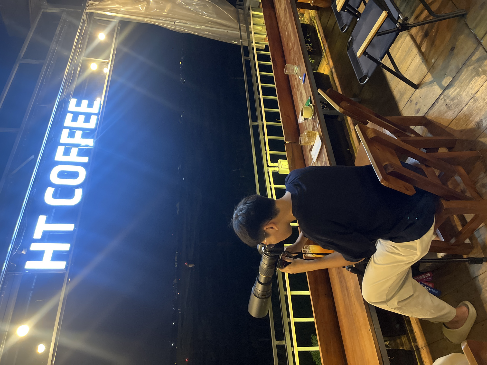 A person is sitting at a wooden table at night, taking a picture with a large camera. The person is wearing a dark t-shirt and light-colored pants. The setting appears to be an outdoor cafe or restaurant with wooden furniture and railings. In the background, there is a large, illuminated sign that reads "HT COFFEE" in bright white letters against a dark sky. There are also other tables and chairs visible, some occupied by other people, and a view of distant city lights. The time of day is night, and the weather appears to be clear. The overall mood seems relaxed and casual, with the person focused on their photography.