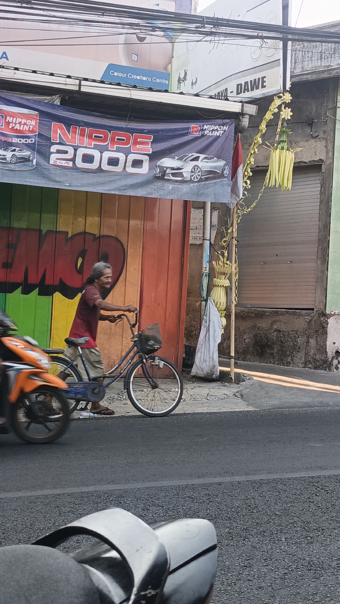 In Dawe, Indonesia, a man with long, graying hair rides a blue bicycle on a paved road. He wears a maroon t-shirt and light-colored shorts. The bicycle has a basket on the front and a dark-colored bag is attached to the handlebars. The man is pushing off the ground with his left foot, suggesting he is in the process of starting to ride or is riding slowly. In the background, a large banner with red and white text reads "NIPPE 2000" and features images of two silver sports cars. The banner is hung above a building with a colorful wall painted with graffiti-like imagery of the letters "EMO" in red, green, yellow, and orange. To the right of the banner, a closed, metal garage door is visible on a building. A few decorative yellow palm frond hangings are suspended from a pole near the garage door. A white plastic bag hangs from the pole below the palm fronds. The foreground shows the edge of a motorcycle. The lighting suggests it is daytime, with sunlight casting shadows on the road.