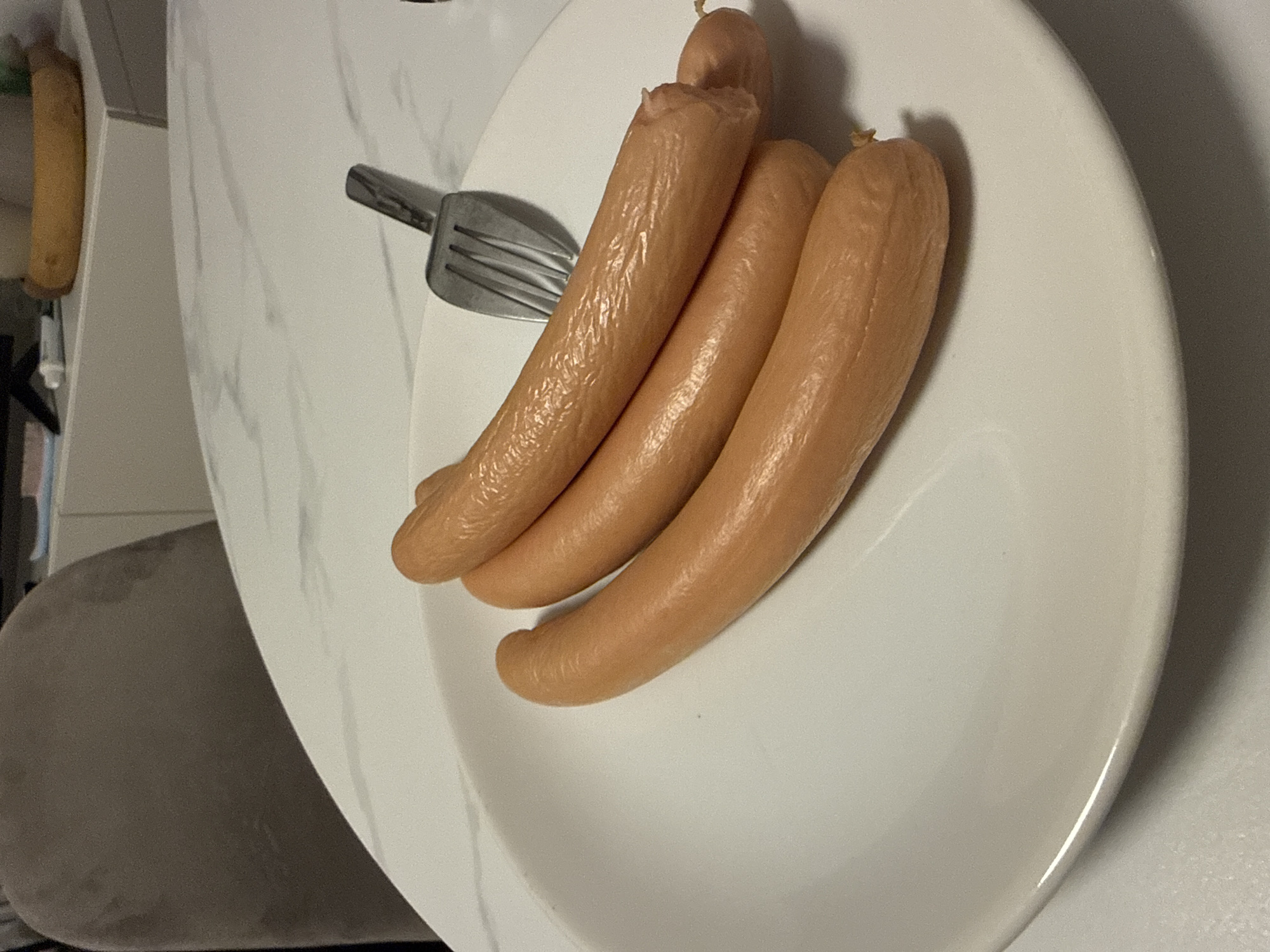 A white plate sits on a surface, holding three cooked sausages and a metal fork. The background is indistinct. Sappada is not discernible in this image.