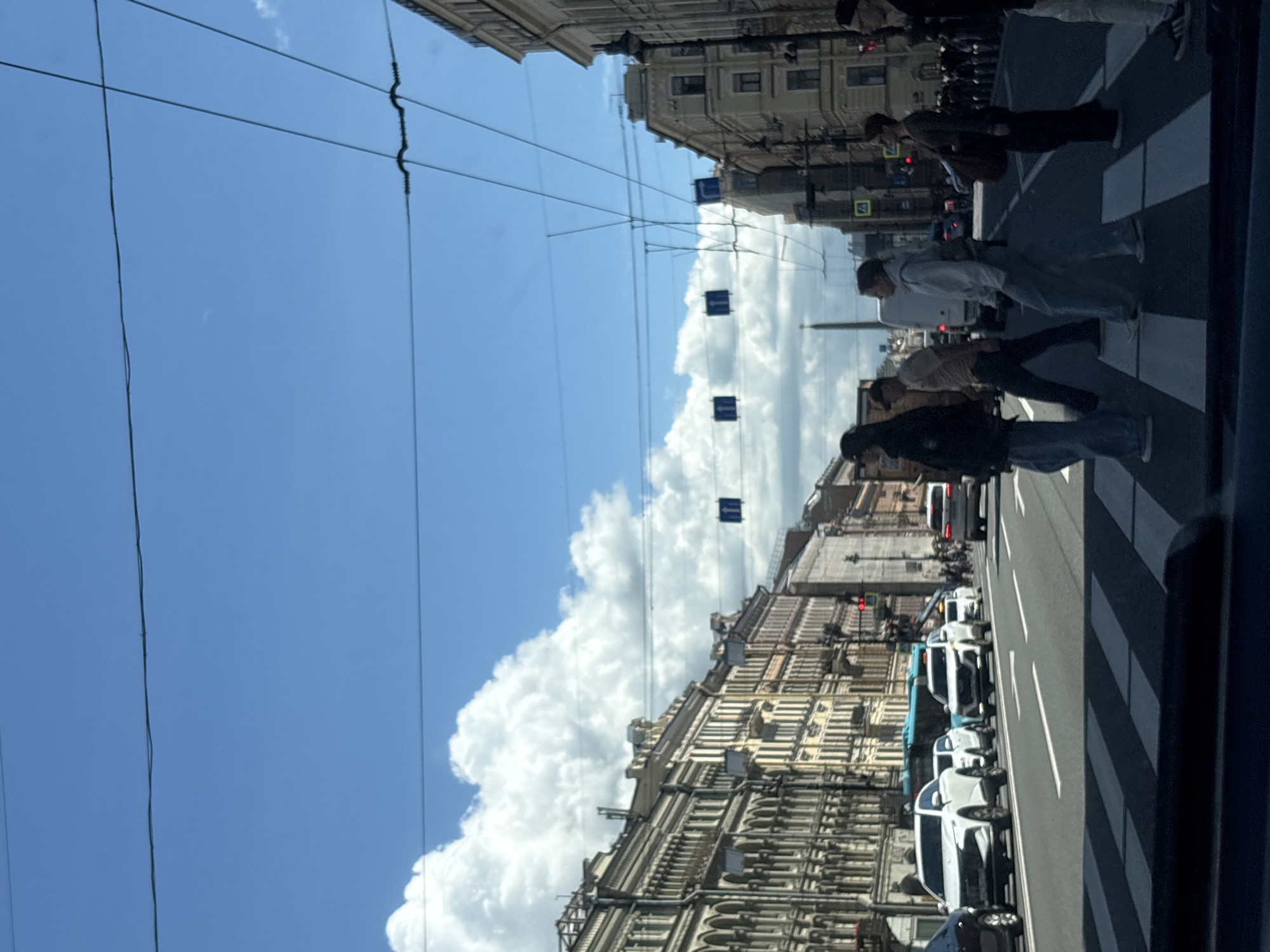 The image captures a street scene in Saint Petersburg, Russia, likely on a sunny day with scattered clouds. The perspective is from a vehicle, showing a wide street with multiple lanes and a pedestrian crosswalk in the foreground.
On the right side of the street, several people are crossing the road at the designated crosswalk. One individual, partially visible on the far right, is wearing white pants and a black shirt. To their left, a woman with short, dark hair and sunglasses is crossing, carrying a large brown bag. Next to her, a man in a light-colored t-shirt and dark pants walks across. Further left, a person in a black hooded jacket and blue jeans is also crossing.
The street is lined with multi-story buildings characterized by classical architecture, featuring intricate facades, numerous windows, and ornamental details. The buildings suggest an urban environment. Traffic is present, with several cars on the road, including white SUVs and a blue bus. Traffic lights and street signs are visible. Overhead, power lines crisscross the sky.
The sky is a clear blue, with large, fluffy white clouds adding visual interest. In the distance, a tall obelisk-like structure is visible, a characteristic feature of some Saint Petersburg landmarks. The overall impression is of a busy urban street in a historic city.
