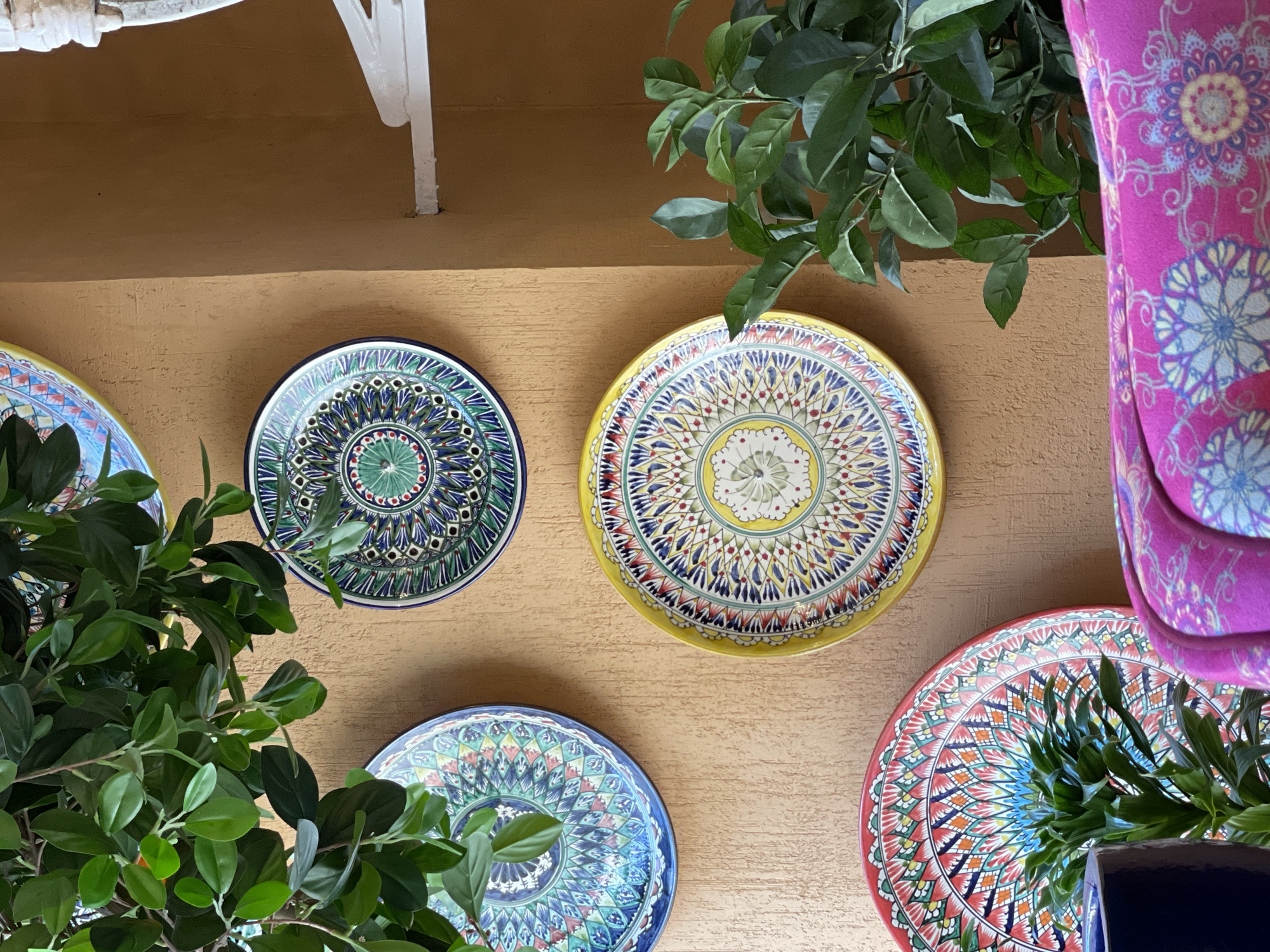 The image shows a wall decorated with four decorative plates, partially obscured by foliage. The plates have intricate, colorful patterns and are arranged seemingly randomly on the wall. The wall itself is a textured beige color. The edges of a white structure are visible at the top left of the frame. A portion of a pink patterned object is on the right edge. The photograph appears to have been taken during the day, judging from the lighting. There are no people visible in the image. There is no information in the image to indicate this is in Saint Petersburg, Russia.
