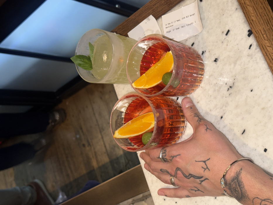 A close-up, high-angle shot depicts three beverages on a light-colored, speckled countertop. Two drinks in clear, ribbed glasses contain an orange-red liquid, each garnished with a triangular orange slice and two green olives. The third drink, in a similar ribbed glass, contains a pale green liquid with a large, clear ice cube and two green basil leaves.
To the left of the two orange-red drinks, a human left hand rests on the countertop. The hand is tattooed with various designs, including a snake-like figure on the back, smaller branch-like patterns, and a detailed eye motif on the forearm, partially visible beneath a thin metallic bracelet with a light-colored stone. A simple silver-toned band ring is worn on the ring finger.
In the blurred background, the lower legs and feet of at least two individuals are visible on a wooden floor, indicating the presence of other people. A dark, possibly reflective, vertical surface is also visible in the background. Two crumpled paper receipts are present on the countertop to the right of the drinks.
The location information visible on one receipt indicates the city of Yerevan, Armenia. Specifically, the receipt prominently displays "Yerevan Mall" (Երևան Մոլ) and lists an address including "Mergelyan 27, Juni Mall, Tigrani 2".
