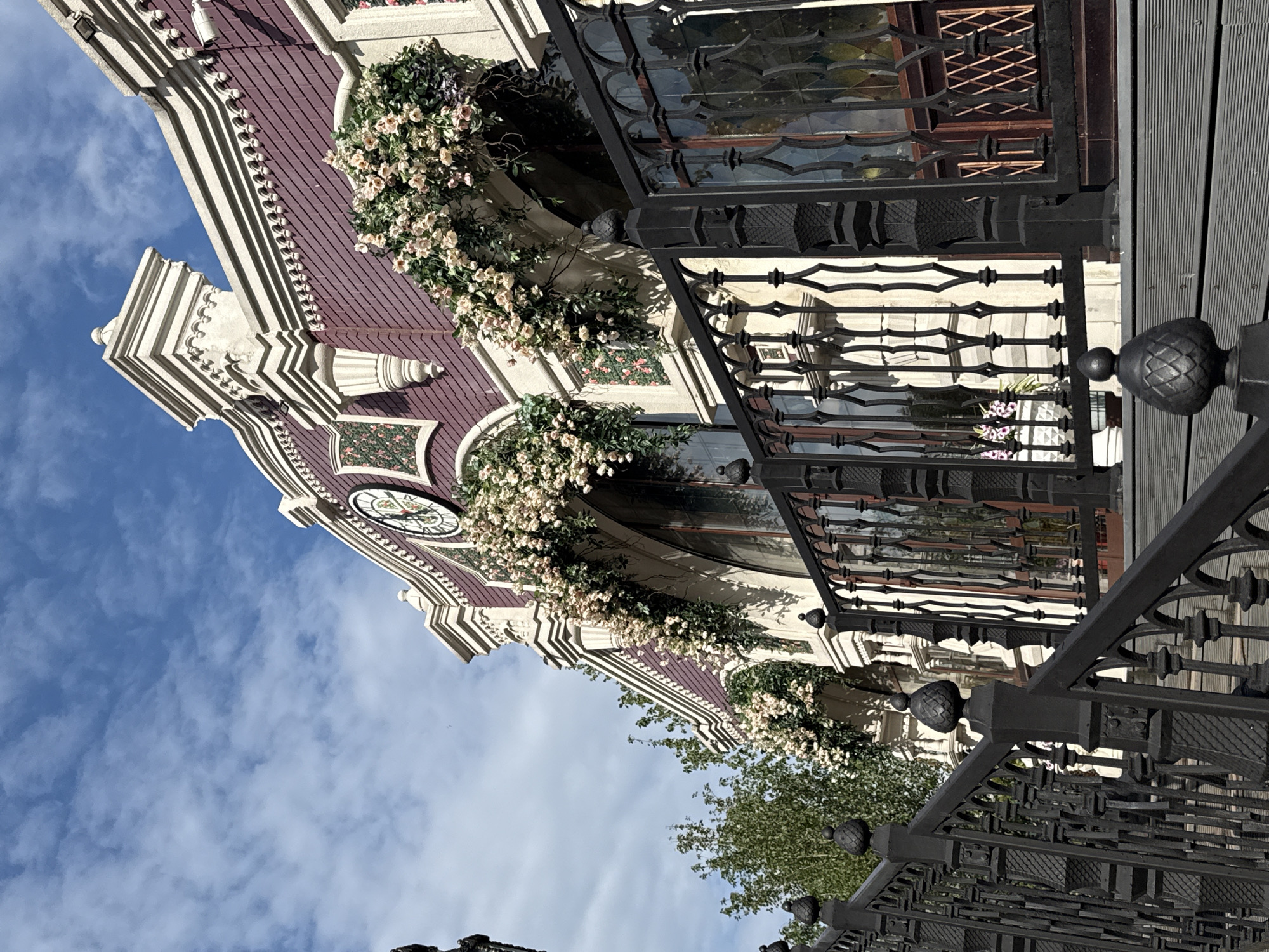 A low-angle, upward-facing shot captures a building facade with ornate architectural details. The building features a maroon roof with decorative white trim and a large, circular clock face with Roman numerals. Cascading floral arrangements, comprised of light-colored blossoms and green foliage, adorn balconies and window ledges. Black wrought-iron railings and decorative elements frame the building and extend into the foreground. The sky above is a bright blue with scattered white clouds, suggesting fair weather during the daytime. Hints of green foliage from trees are visible at the lower right and middle of the frame. The architectural style and decorative elements suggest a historical or European-inspired setting, consistent with the provided location context of Moscow, Russia. No people are visible in the frame.