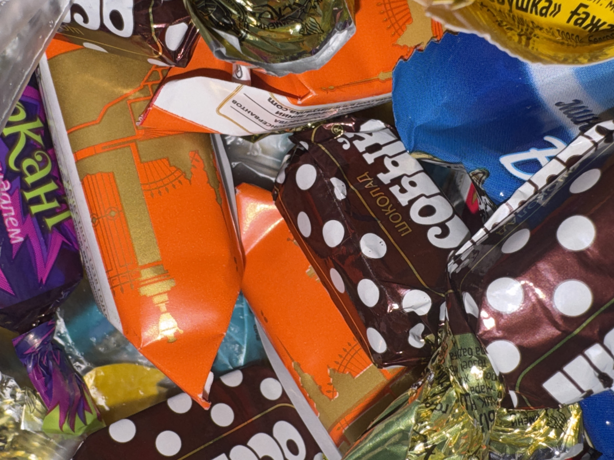 This is a close-up, overhead shot of a pile of assorted candy wrappers. The wrappers are a mix of vibrant colors, including orange, purple, blue, gold, and brown, with some featuring polka dot patterns. Some of the wrappers appear to be made of foil, while others are paper.
Visible text on the wrappers includes Cyrillic script. Some of the text is partially obscured, but visible words and phrases include "ШОКОЛАД," "СОБЫЙ," "Кант," and a website address "yanka.com". There are also phrases in Russian that may indicate ingredients or brand names, such as "ИНСЕРВАНТОВ" and "же «". The text suggests the candies are likely chocolates and from a Russian or Belarusian manufacturer.
The scene is a chaotic jumble of sweets, suggesting they might have been recently opened or are being stored together. There are no people or animals visible in the image. The lighting is bright, indicating good illumination, possibly from an indoor setting. The overall impression is one of a sweet treat assortment, with a focus on the colorful and varied packaging. No specific time of day, weather, or emotional cues are discernible from the image. The location context provided, "Яромінскі сельскі Савет, Belarus," suggests the origin or a possible location of these candies.