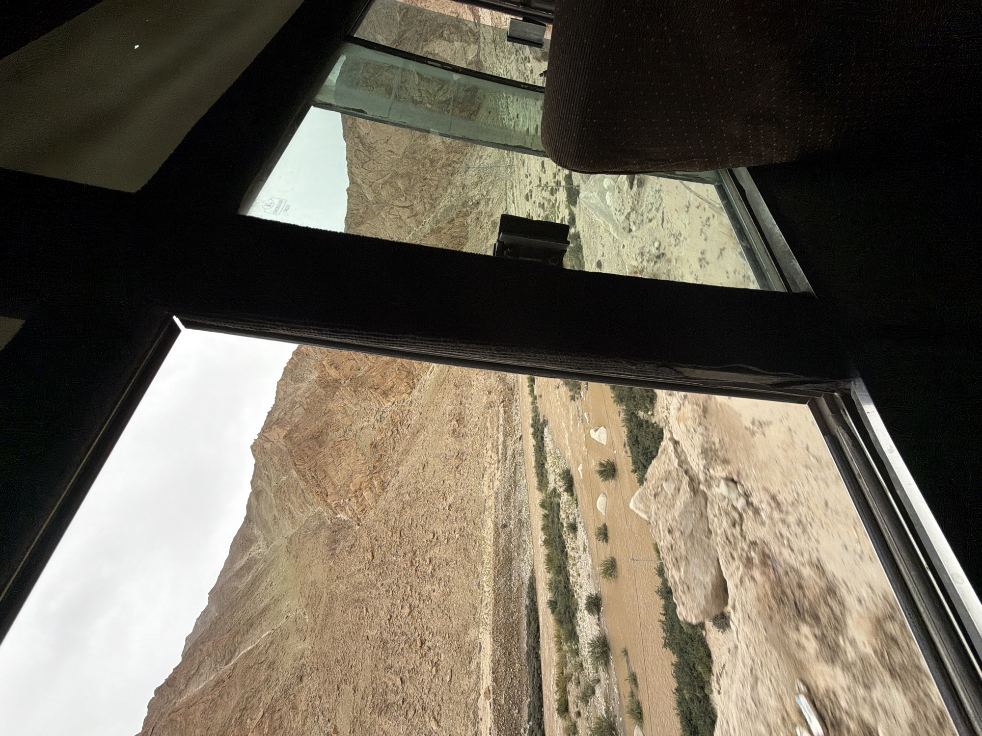 The image depicts a view from inside a vehicle looking out at a mountainous, arid landscape. The foreground shows the interior of the vehicle with dark brown seats and part of a window frame. Through the window, a wide, shallow, muddy river flows through a valley. The riverbanks are lined with sparse green vegetation and scattered rocks. Beyond the river, the terrain rises into steep, rocky, beige-colored mountains. The sky is overcast and a light gray. The location is identified as Hymia, India.