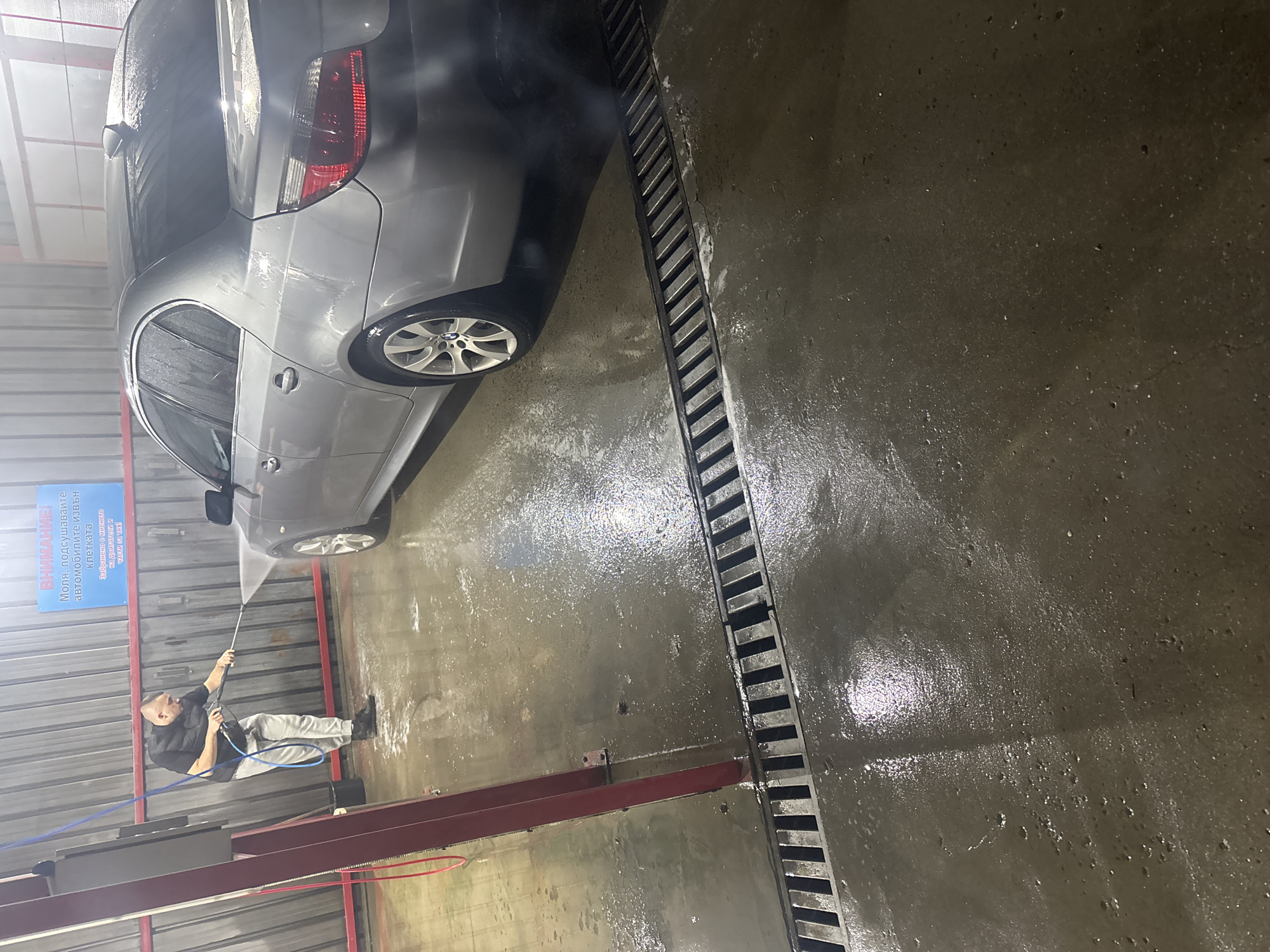 The image shows a car wash. A silver sedan is partially visible at the top left, being washed by a man with a pressure washer. The man is wearing a black vest, gray pants, and black shoes. He is holding the pressure washer with both hands, directing the spray towards the car. To the right of the car is a grated drainage system running across the floor. The floor is wet and made of concrete. Above the man, a blue sign is visible with text in Bulgarian. A red support structure with tubing is also seen in the lower-left portion of the image. The overall scene suggests an indoor car wash facility. The Bulgarian text on the sign indicates the location could be in Bulgaria, and based on the OCR, it reads "ВНИМАНИЕ! Моля, подсушавайте автомобилите извън клетката. Забранено е миенето на двигатели и части за тях!" which can be translated as "Attention! Please dry the cars outside the cell. It is forbidden to wash engines and parts for them!" The location, Plovdiv, Bulgaria, is not directly identifiable in the image.