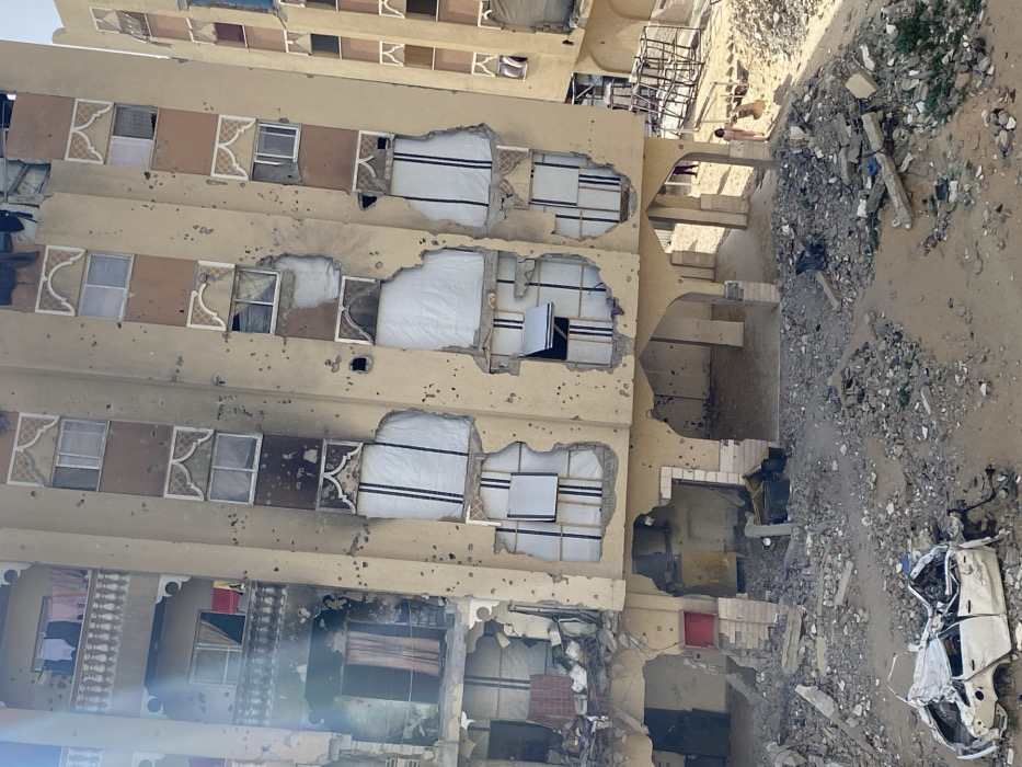The image depicts a multi-story residential building in Khan Yunis, Palestinian Territory, exhibiting extensive damage. The building's facade, primarily light beige, is riddled with numerous impact marks consistent with bullet or shrapnel hits. Large sections of the external walls and window areas are destroyed, exposing interior rooms. Many windows are shattered or entirely absent, with some openings covered by white tarpaulin or makeshift panels. The left side of the building shows more severe structural compromise and charring, suggesting fire damage. A balcony on an upper floor on the left still has clothes hanging. The ground floor features several arched entryways leading to open, rubble-strewn spaces.
In the foreground, a heavily damaged white sedan, crushed and partially overturned, lies amidst a large field of debris composed of broken concrete, twisted metal, and other construction materials. The ground surface is predominantly sandy dirt. To the right of the debris field, a single individual, possibly a child, dressed in light attire, stands on the sandy ground facing away from the viewer. Another less distinct figure is partially visible further back beneath an archway on the building's right side. The scene is illuminated by bright daylight.