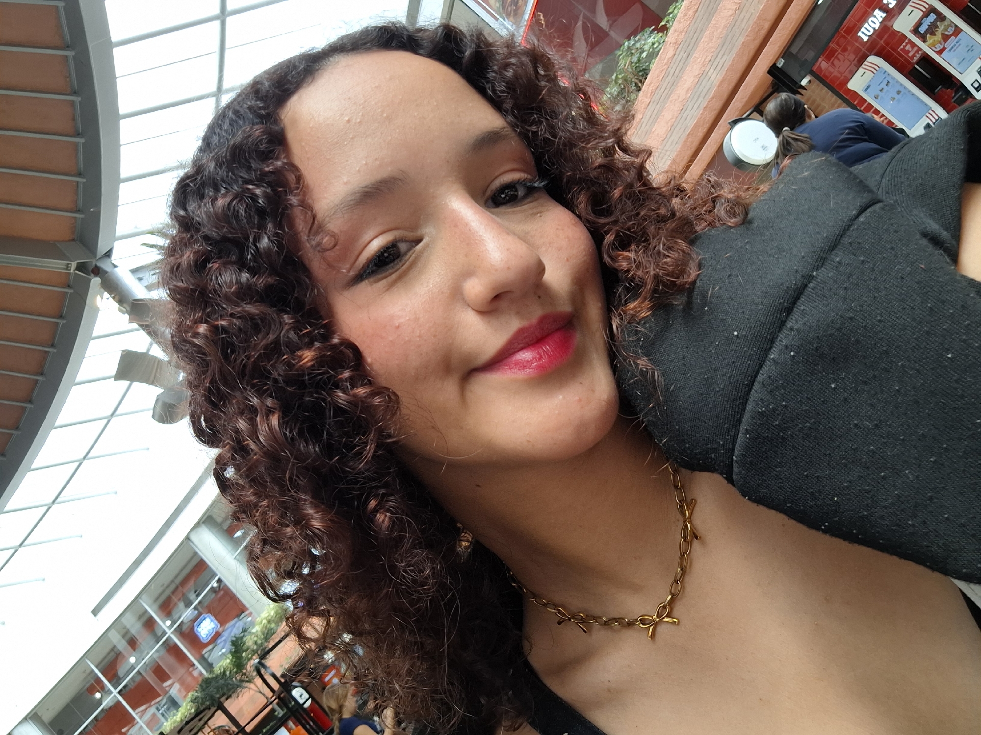 The image is a close-up selfie of a young woman with curly, dark brown hair, taken indoors. She is wearing a black jacket over a dark top and a gold chain necklace. Her head is tilted, and she is smiling slightly with bright red lipstick. The background suggests a public space with modern architecture, possibly a shopping mall or a large indoor atrium, characterized by a curved ceiling with visible beams and large windows. In the blurred background, there are hints of shops with red signage and displays, and other people are visible, one wearing a blue shirt and another with dark hair. The overall lighting is bright, indicating natural light coming from the windows. The location is identified as the city of Pampatar, Venezuela.