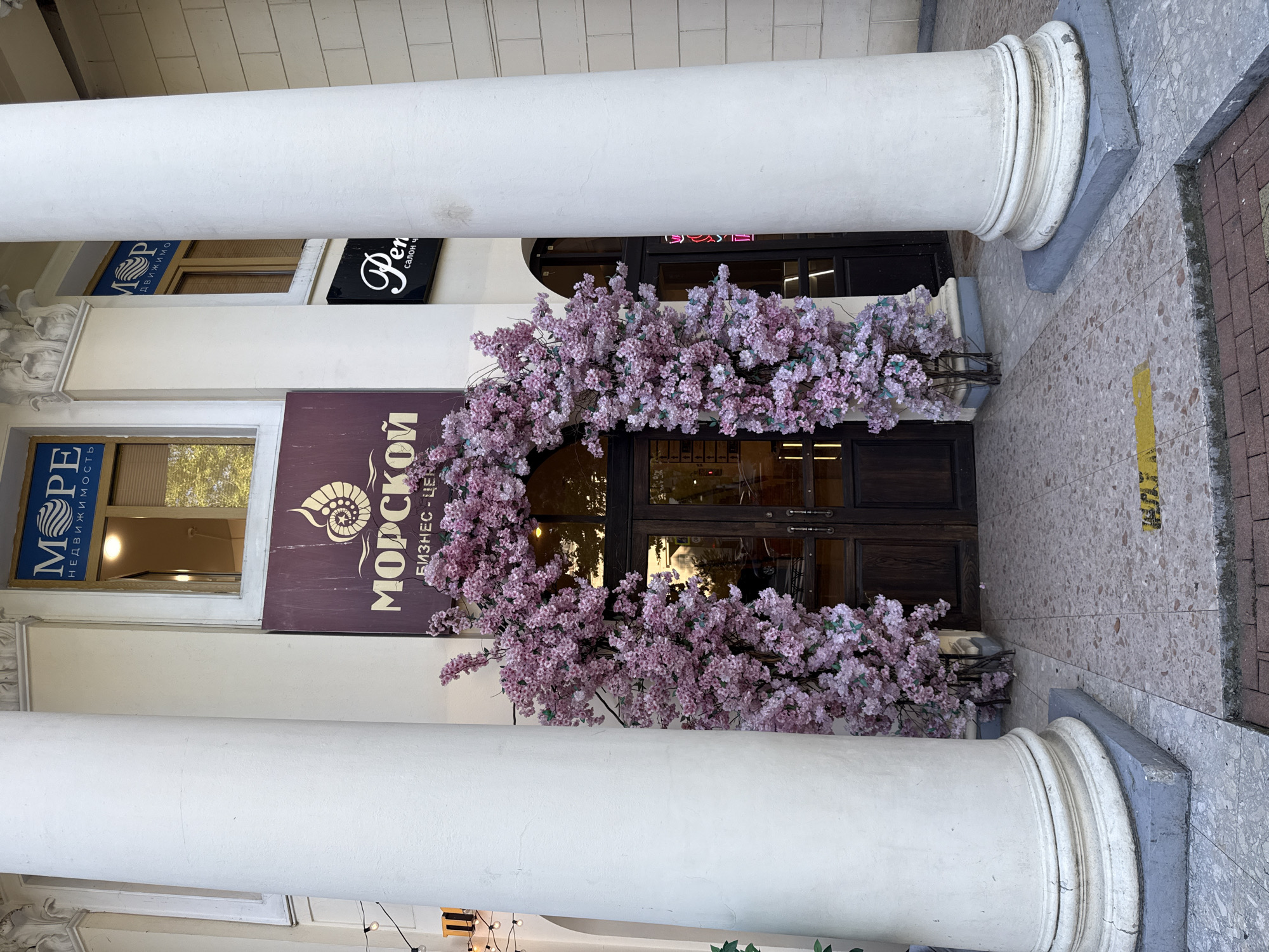 An exterior, eye-level, full shot captures the entrance to a business center in Sochi, Russia. Two large white columns flank a dark wood door, framed by an archway adorned with artificial pink cherry blossoms. Above the door, a sign reads "МОРСКОЙ БИЗНЕС-ЦЕНТР" (MORSKOY BUSINESS CENTER) with a seashell logo. To the right of this sign, a smaller black sign with white lettering says "Pen" and "салон ч" (salon ch). Above the "МОРСКОЙ" sign, a blue sign with white lettering states "МОРЕ НЕДВИЖИМОСТЬ" (MORE REALTY). The ground is paved with light-colored speckled tiles, with a yellow line painted on the ground in front of the entrance. The lighting suggests it is daytime, with a slightly overcast sky. No people are visible in the frame.