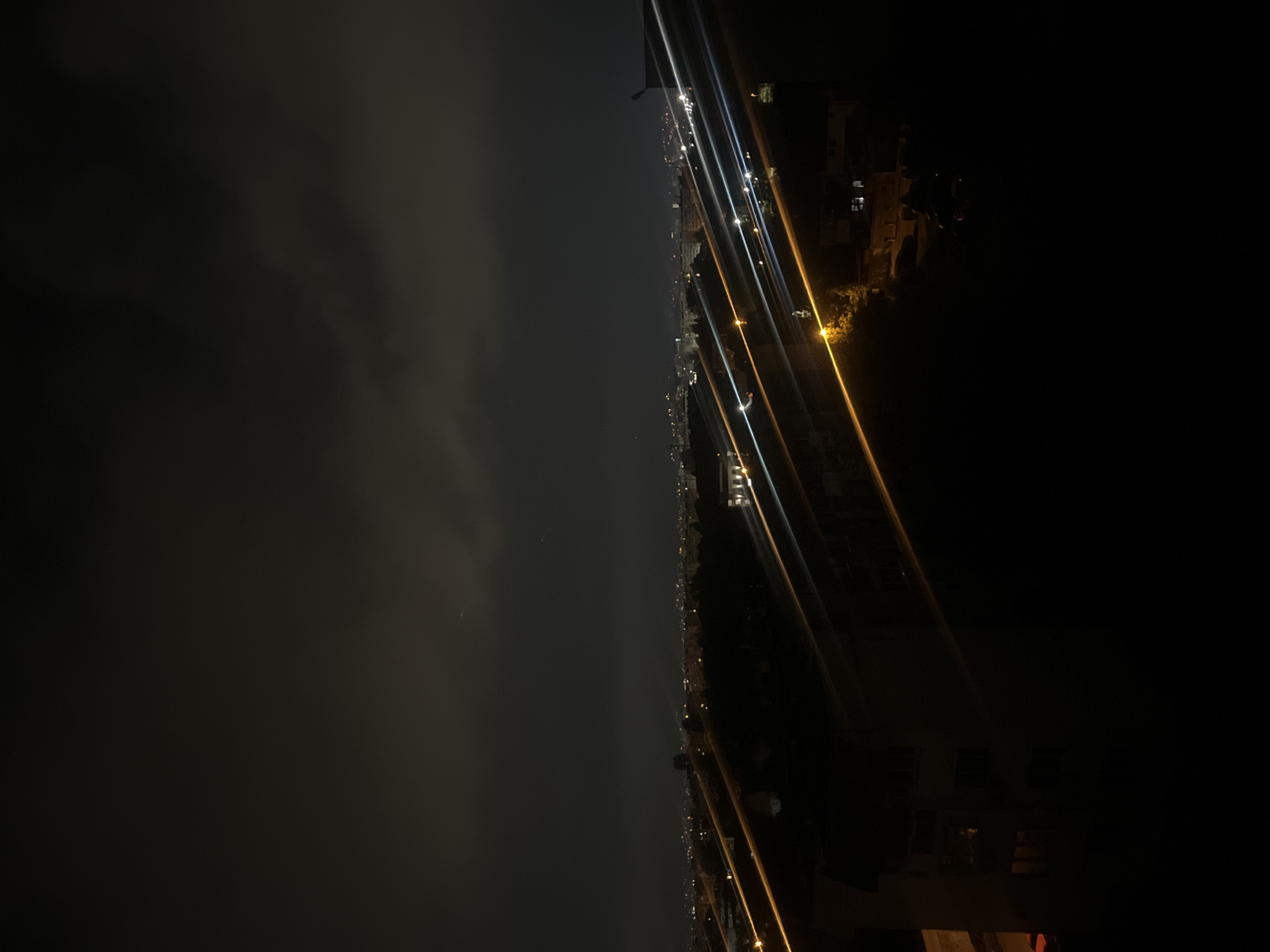 At night, a vertically oriented cityscape is split between sky and city. On the left, a dark, cloudy sky occupies half of the image. A faint, crescent moon is barely visible. On the right, the cityscape is defined by lines of streetlights and building lights, creating streaks due to the camera's movement during the long exposure. The lights give the impression of a densely populated urban area. In the lower right corner, the edge of a structure is visible. The image's low light and indistinct features make specific location identification challenging, but the arrangement of the scene resembles an overview of part of Sofia, Bulgaria.