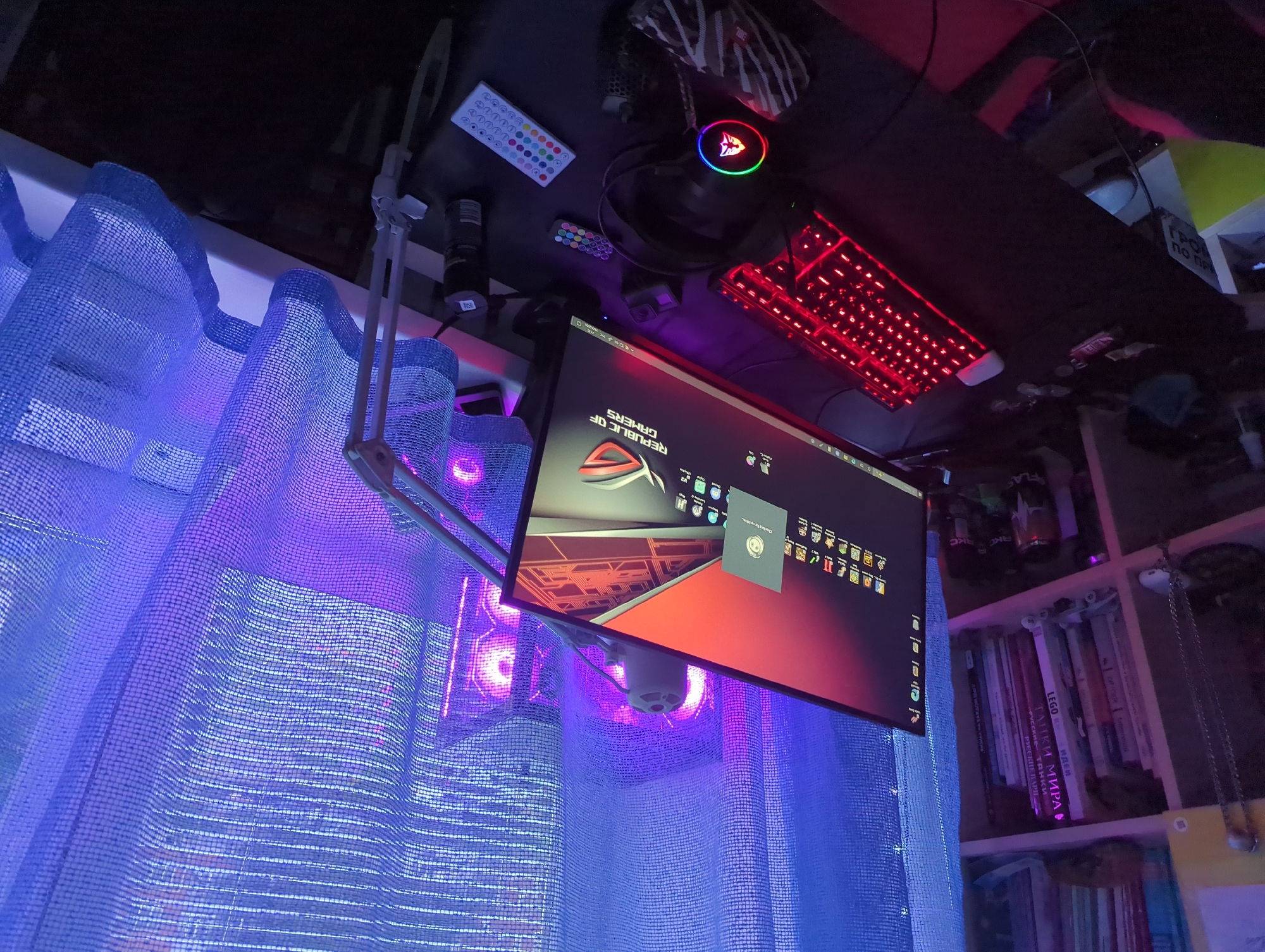 The image shows a gaming setup in what appears to be a bedroom. A large computer monitor displays a gaming desktop with the "Republic of Gamers" logo visible. A red-backlit keyboard is in front of the monitor, and to its right, a black headset with a glowing RGB logo rests on the desk. Several remote controls with colorful buttons are also on the desk, along with a black spray can and a GoPro camera. A bookshelf filled with books is visible to the left of the desk, and a blue, translucent curtain is behind the computer, illuminated by purple and blue LED lights, which also light up the computer tower visible through the curtain. The overall scene suggests a personal gaming space, likely within a residential setting. There is no information in the image that can confirm or deny the location in the city of Votkinsk, Russia.