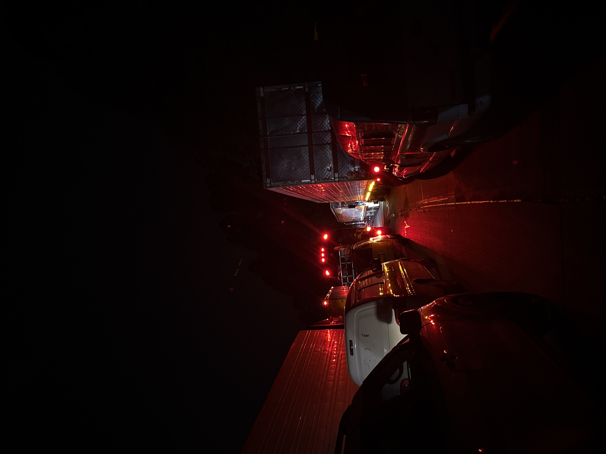 The image depicts a nighttime scene featuring a line of vehicles on what appears to be a road or highway, viewed from inside another vehicle. The scene is dominated by darkness, illuminated primarily by the red taillights and marker lights of the vehicles.
In the foreground, the side and rear of a vehicle, possibly a car or SUV, with a white body panel is visible on the right. Its red taillight casts a glow on the adjacent surface. Behind this vehicle, a large semi-trailer truck is prominently featured, seen from its side. Its dark, possibly insulated or quilted, silver-gray trailer is lined with multiple glowing red marker lights. Further down the road, an array of red lights from additional vehicles extends into the distance, suggesting a long line of traffic. The road surface below reflects the red lights, indicating it might be wet or reflective asphalt.
The activity taking place appears to be a traffic jam or slow-moving convoy, as indicated by the illuminated brake lights and the stationary appearance of the vehicles. The time of day is clearly night, with no natural light visible. There are no discernible people or visible text in the image. No specific weather conditions are evident, though reflections suggest a potentially wet surface. The provided location context for this scene is San Marcos Huixtoco, Mexico.
