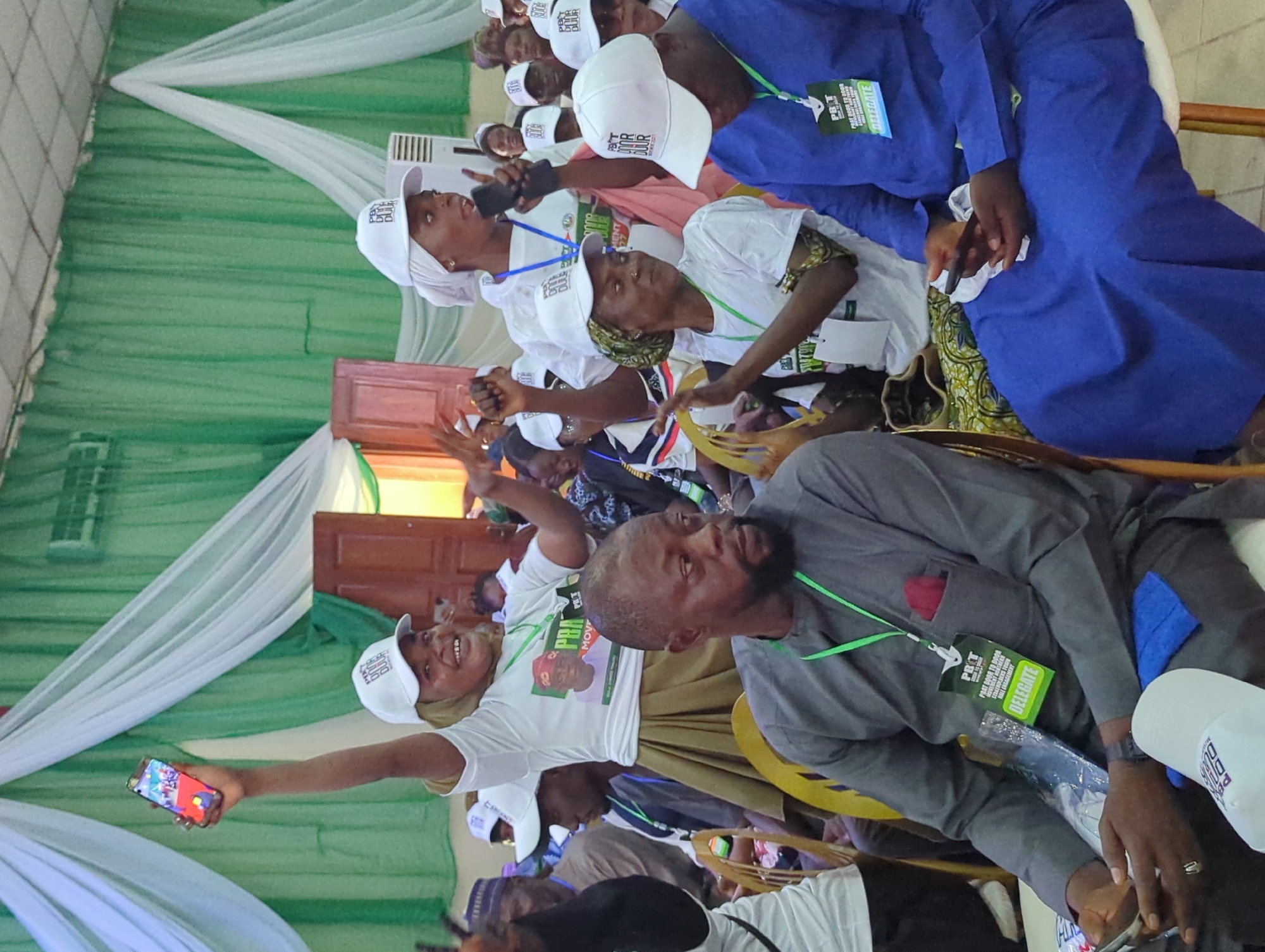 Here is a forensic description of the image:
**Scene:** An indoor gathering of people, likely a political event based on the campaign-related attire.
**People:** There are multiple people in the image. Most of them are wearing white caps with the letters "PBT" on them, along with lanyards that also have "PBT" and "DELEGATE" printed on them. One person is holding up a mobile phone, seemingly taking a selfie. Another person, wearing a grey shirt, appears to be seated and is looking off to the side. Some individuals are dressed in traditional attire, including blue robes.
**Objects:** The objects visible include chairs, white and green drapes in the background, a wooden door, mobile phones, lanyards, and campaign apparel.
**Actions:** People are either seated, standing, talking, taking photos, or observing. The general action suggests participation in the event.
**Interactions:** There is a sense of community and collective action. People are engaging with each other, possibly discussing matters related to the event's purpose.
**Environment:** The location seems to be a hall or large room decorated for an event.