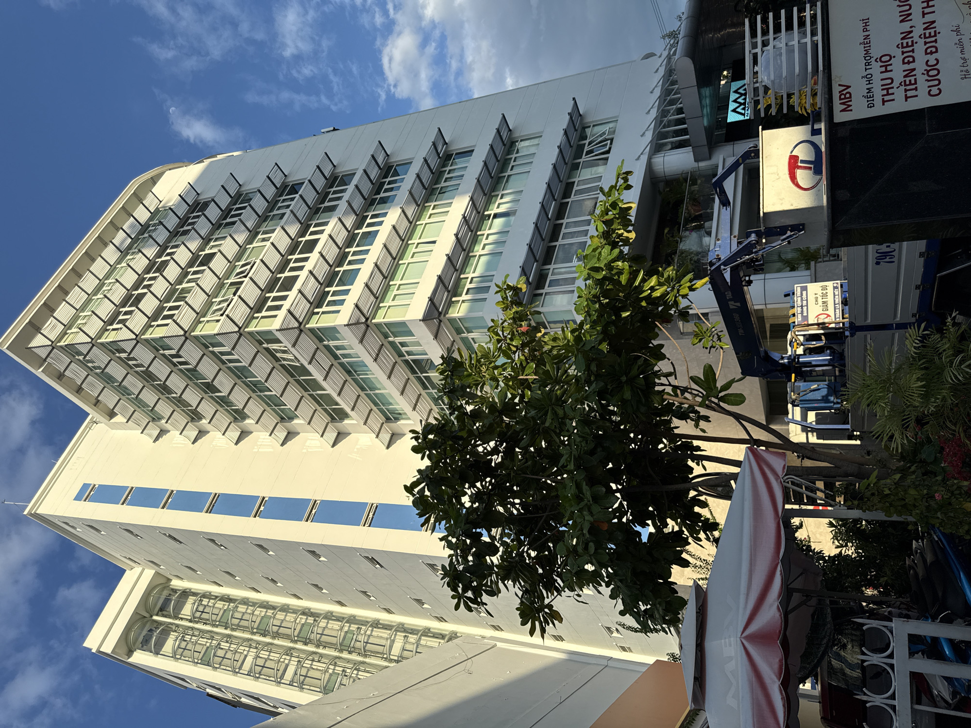 The image depicts a modern, multi-story white building with a distinctive facade featuring numerous horizontal sunshade elements above the windows. To the left of this building, another section of a similar structure is visible, distinguished by transparent cylindrical elevator shafts. In the foreground, a large tree with dense green foliage partially obscures the lower floors of the buildings. A white umbrella with red trim is set up in front of the buildings, and beyond it, parts of what appears to be construction equipment, including a blue crane on a truck, are visible. Signs with Vietnamese text are present, including one that reads "MBV" and another that seems to be an advertisement for services. The sky above is a clear blue with scattered white clouds. The scene is likely a street-level view in an urban area of Nha Trang, Vietnam, given the architectural style and the presence of Vietnamese signage.