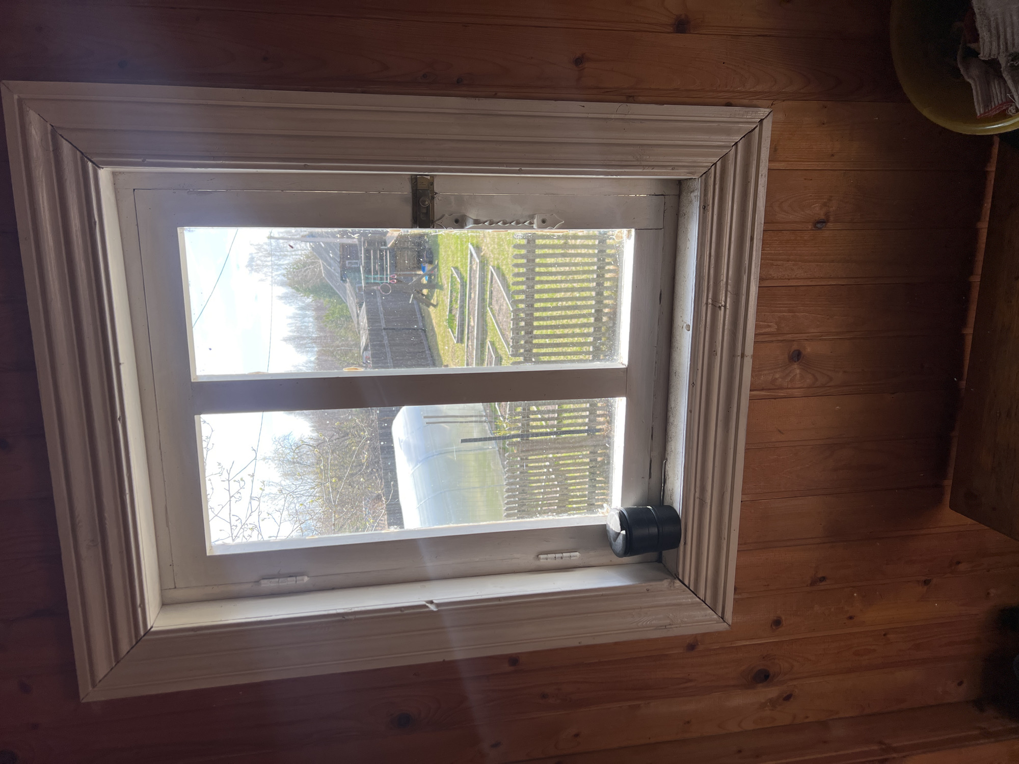 The image shows a window in a wooden building. The window is white with a decorative frame. Outside the window, there is a view of a yard with a greenhouse, a picket fence, and some garden beds. The location is specified as Чернышово, Russia. A black cylindrical object is attached to the bottom right of the window frame.