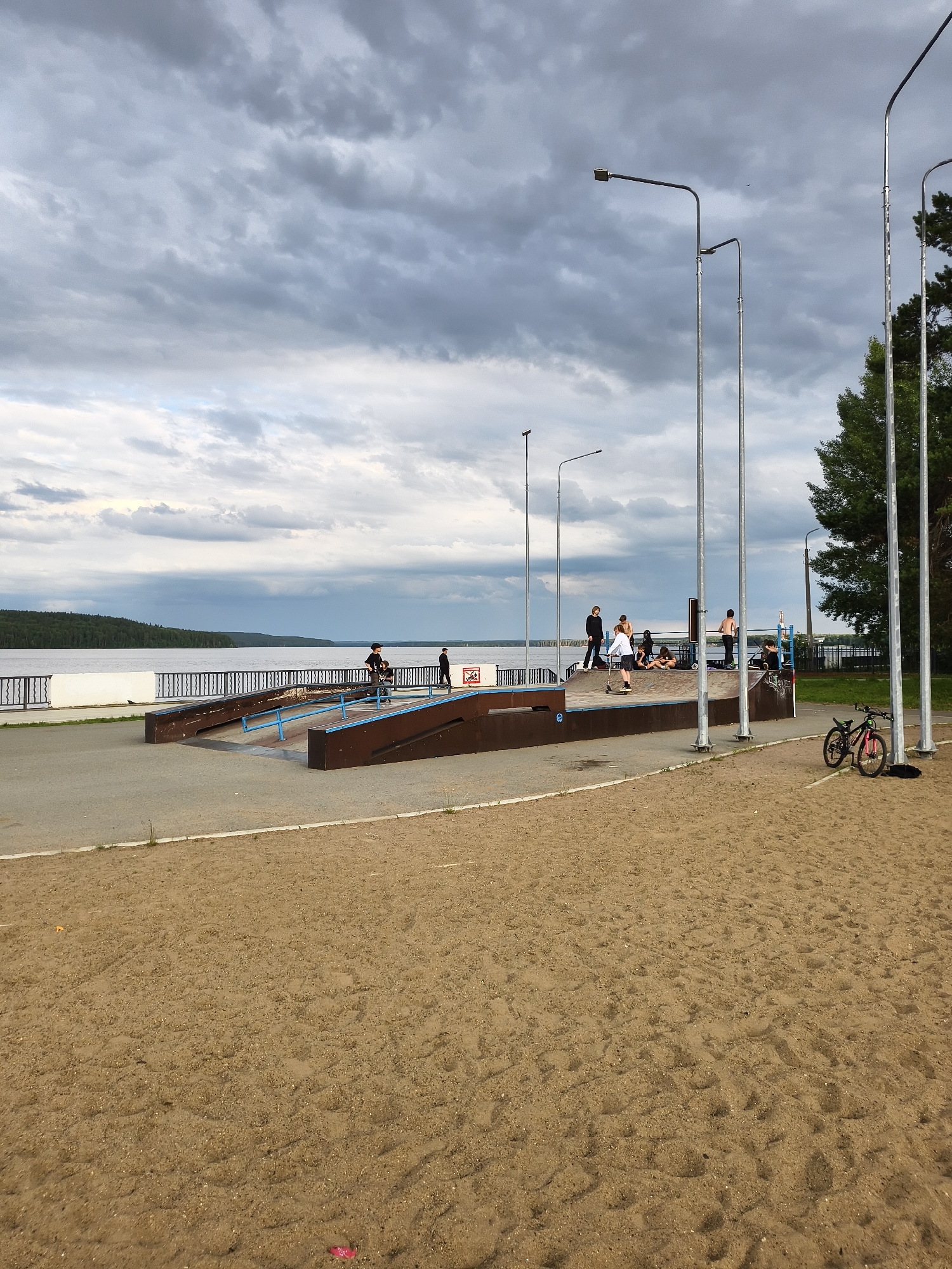 The image captures a skatepark situated near a large body of water, likely a river or lake, with a wooded shoreline in the distance. The sky is overcast with dramatic, dark grey clouds, suggesting impending rain or a storm. The time of day appears to be late afternoon or early evening, given the diffused light.
Several people, mostly young individuals, are present at the skatepark. Some are actively skateboarding or on scooters, while others are gathered, watching or resting. One person is on a scooter on a ramp, while others are positioned on and around the skatepark structures. The skatepark itself is made of concrete and wood, with various ramps and ledges.
In the foreground, a sandy area is visible, with tire tracks from bicycles suggesting recent activity. Two bicycles are parked near the skatepark; one is black and red, and the other is a lighter color. In the background, a white barrier with a dark railing separates the skatepark area from the water. Tall lampposts are spaced along the paved area, indicating the space is likely used in the evening. The overall mood is casual and recreational, despite the somber weather. No discernible text is visible in the image. The location is identified as Chaikovsky, Russia, which aligns with the presence of a large body of water and forested landscape.