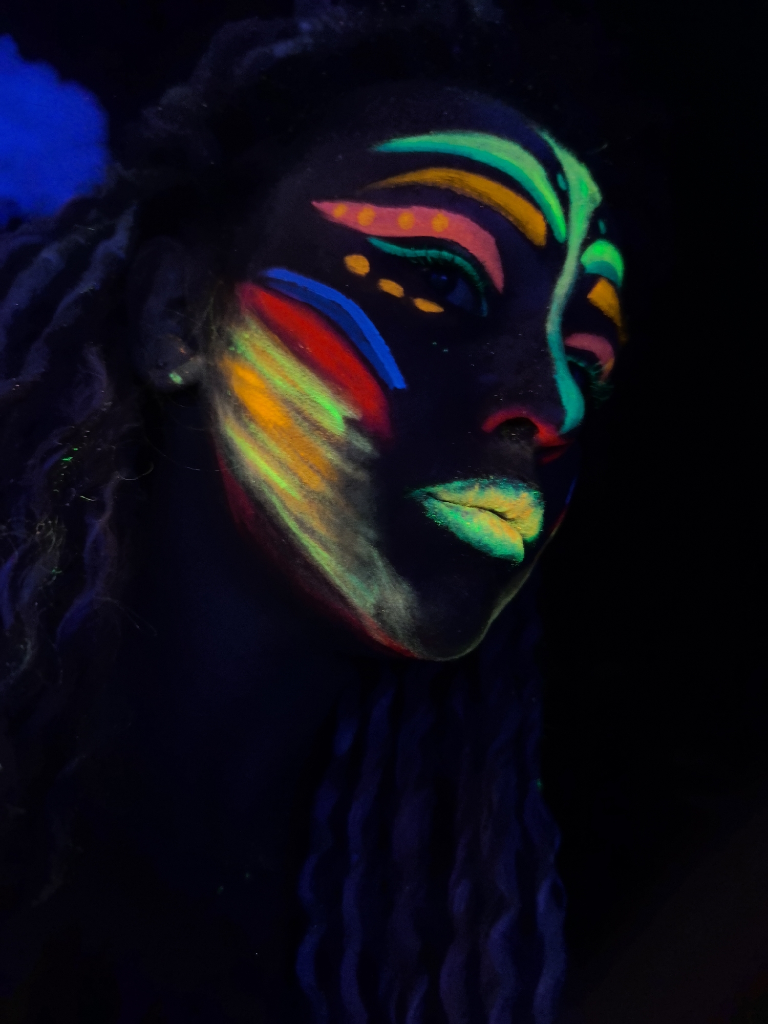 A low-angle shot of a person with dark skin under ultraviolet light, revealing elaborate face paint. The paint features vibrant neon colors like green, yellow, pink, red, and blue, arranged in geometric patterns around the eyes, cheeks, and lips. The individual has dark dreadlocks. The background is dark, emphasizing the glowing paint. The scene is unlikely to be in the city of Noviy, Russia; the photo is likely taken in a studio.