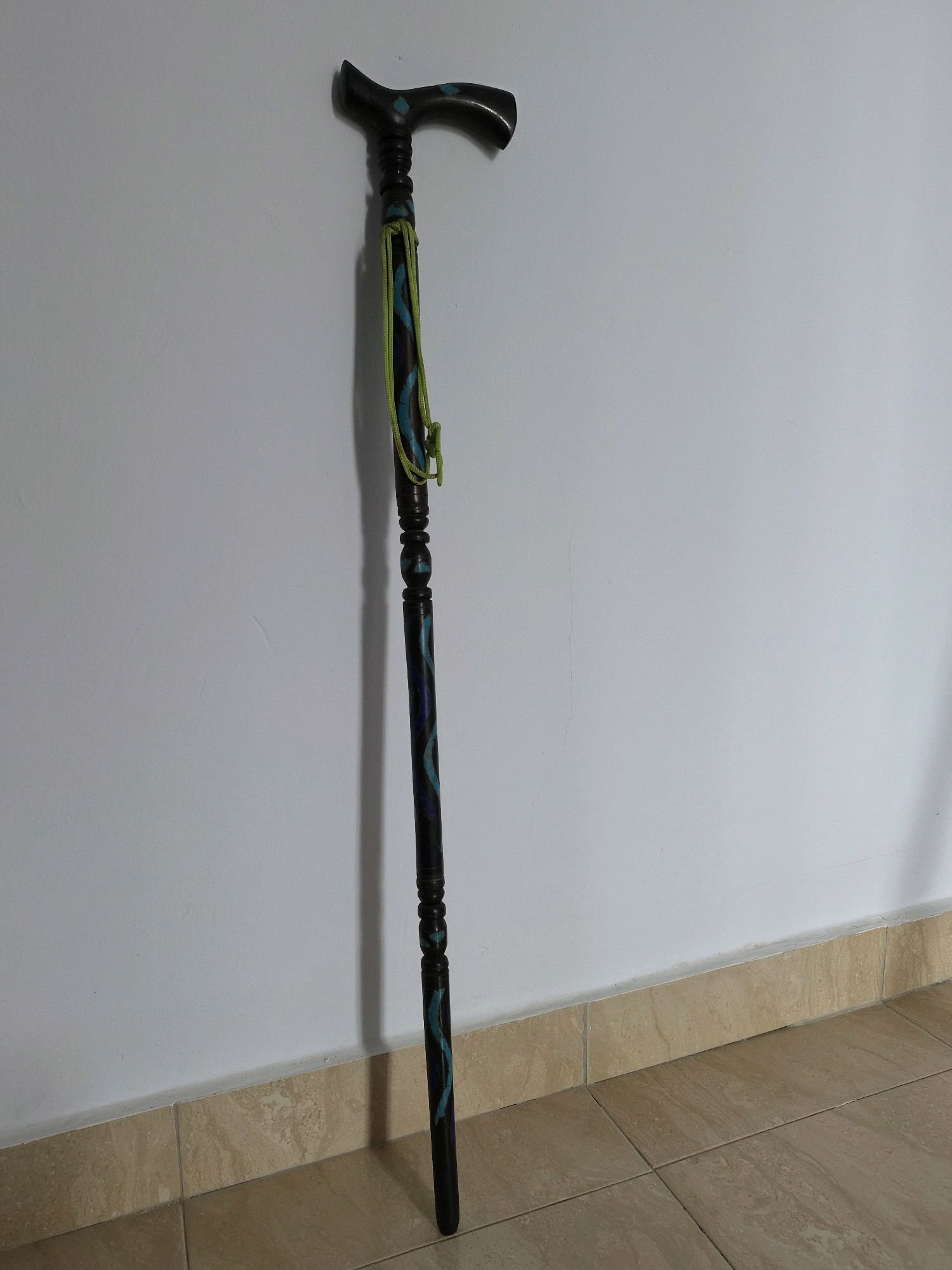 A dark wooden cane with decorative blue and green swirling patterns stands upright against a white wall, leaning slightly to the right. The cane has a curved handle and a twisted rope attached near the top. The base of the cane rests on a tiled floor composed of beige, brown, and tan square tiles. The wall appears to be painted, with some minor scuffs and cracks visible. The lighting suggests an indoor environment, possibly a room in a residential setting. The specific location within Srimahi, Indonesia, cannot be determined from the visual information provided.