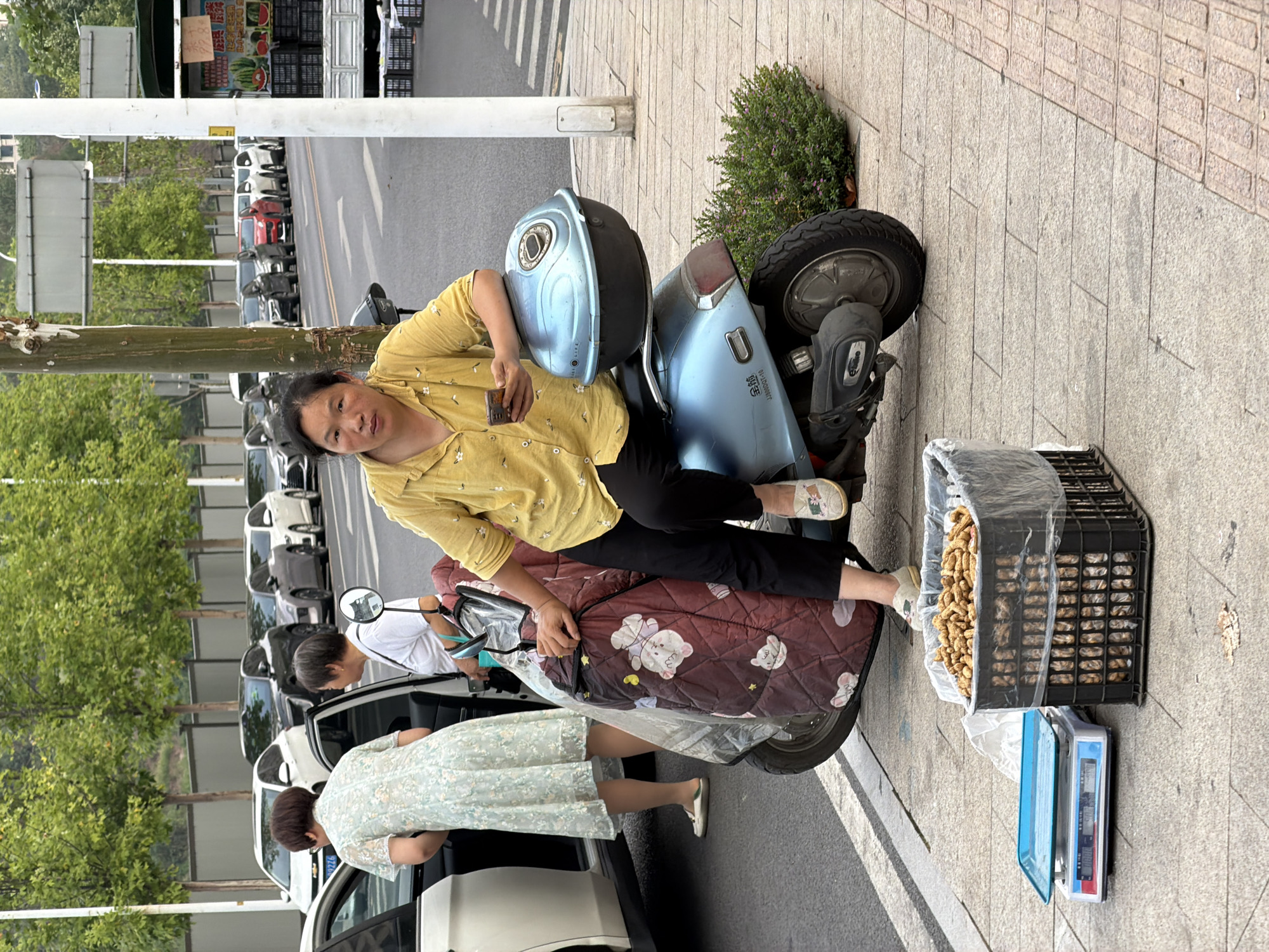 In Jiangjin, China, a woman in a yellow shirt sits on a blue scooter with a black and blue storage box. She holds a mobile phone and appears to be looking at it. In front of the scooter, a black crate is filled with peanuts, and next to it, a weighing scale sits on the sidewalk. Two other women are visible near a white car, one in a light blue floral dress and the other in a white shirt. The background shows a street with parked cars and trees.