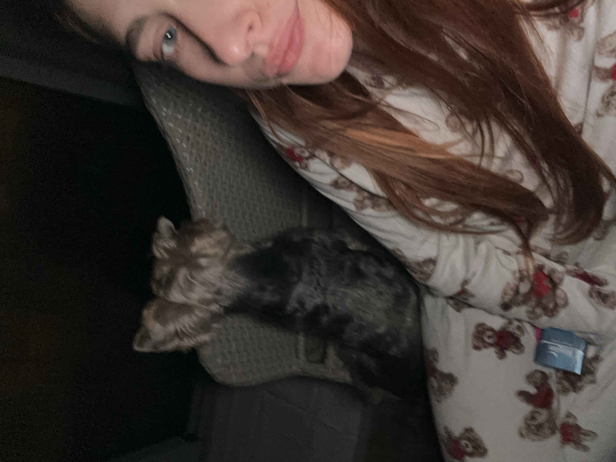 A woman with long reddish-brown hair and fair skin is lying at an angle in a chair. She is wearing pajamas patterned with teddy bears. Next to her and partly obscured by the chair is a small dog with shaggy fur. The background includes a dark area, possibly a doorway or shadowed space. No information indicates this is in Budva, Montenegro.